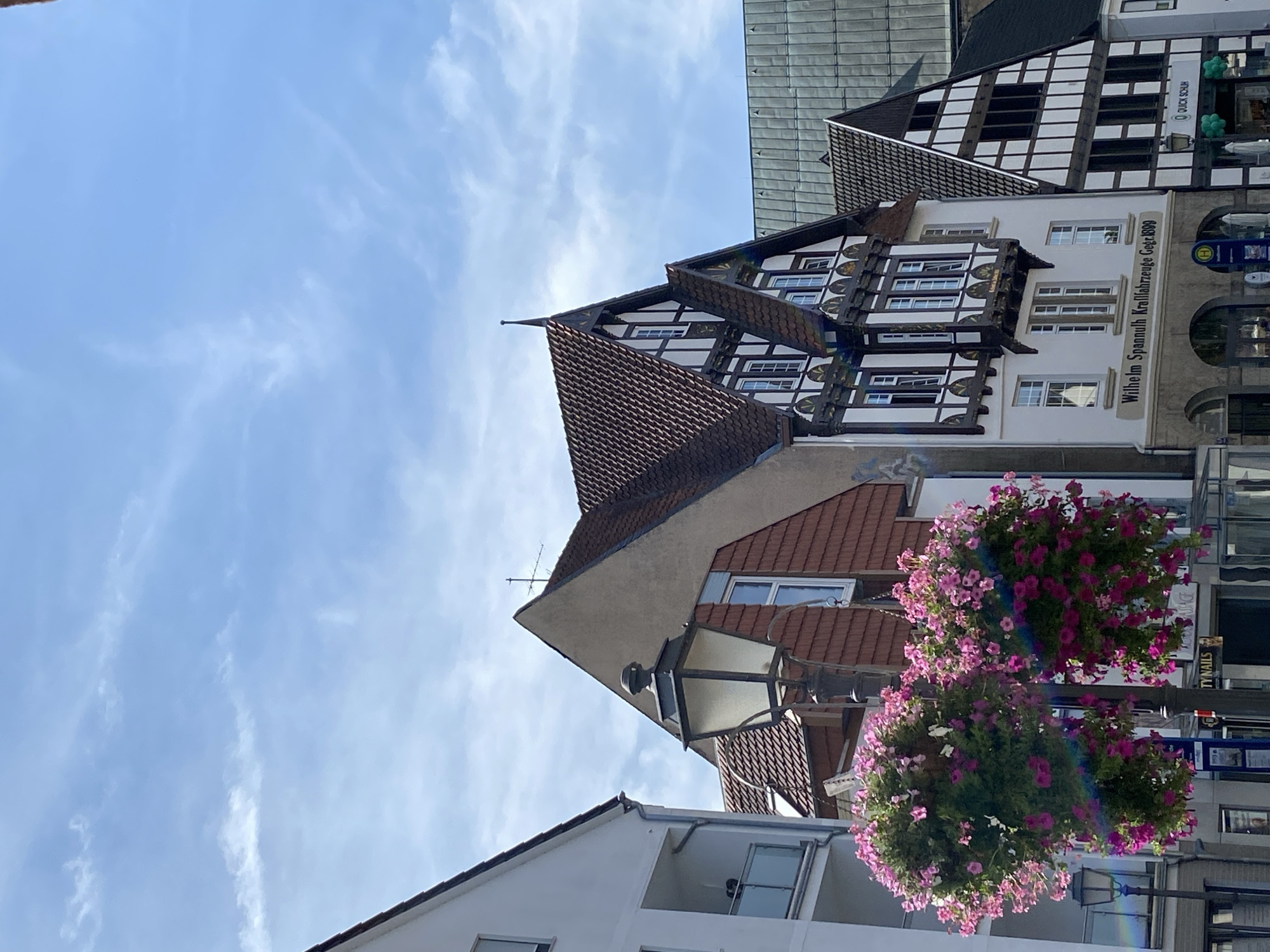 In Bückeburg, Germany, a street view displays traditional half-timbered buildings against a bright blue sky with wispy clouds. A lamppost with two large hanging baskets of pink petunias stands in the foreground. To the left, a modern white building with a balcony is visible. The central building features a prominent gable, dark timber framing, and white stucco, with a sign that reads "Wilhelm Spannuth Kraftfahrzeuge Gegr. 1899." To its right, another half-timbered building with a similarly steep roof is partially visible, with a sign for "QUICK SCHUH." The ground level of the buildings appears to house shops, indicated by awnings and signage. The overall scene depicts a charming European streetscape on a sunny day.