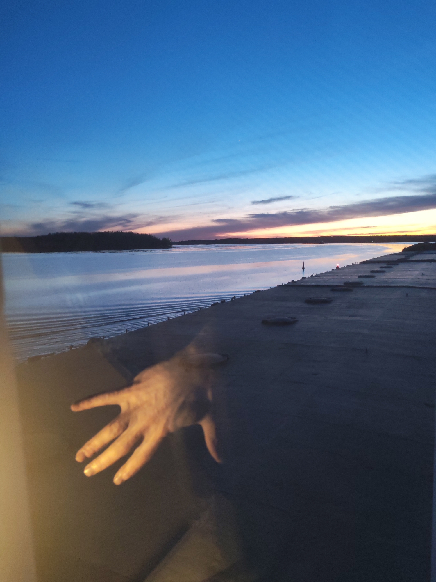 A view from inside a structure or vehicle, possibly a window or porthole, looks out over a body of water reflecting the colors of a sunset or twilight. The sky transitions from a deep blue at the top to hues of orange, pink, and purple near the horizon. A dark landmass or island is visible in the distance. Along the edge of the water, there is a man-made structure with evenly spaced, dark, circular or oval elements. A semi-transparent human hand is superimposed in the foreground, near the bottom left, suggesting the viewer is looking through a window or barrier. The image is shot in low light. The location is Topornya, Russia.
