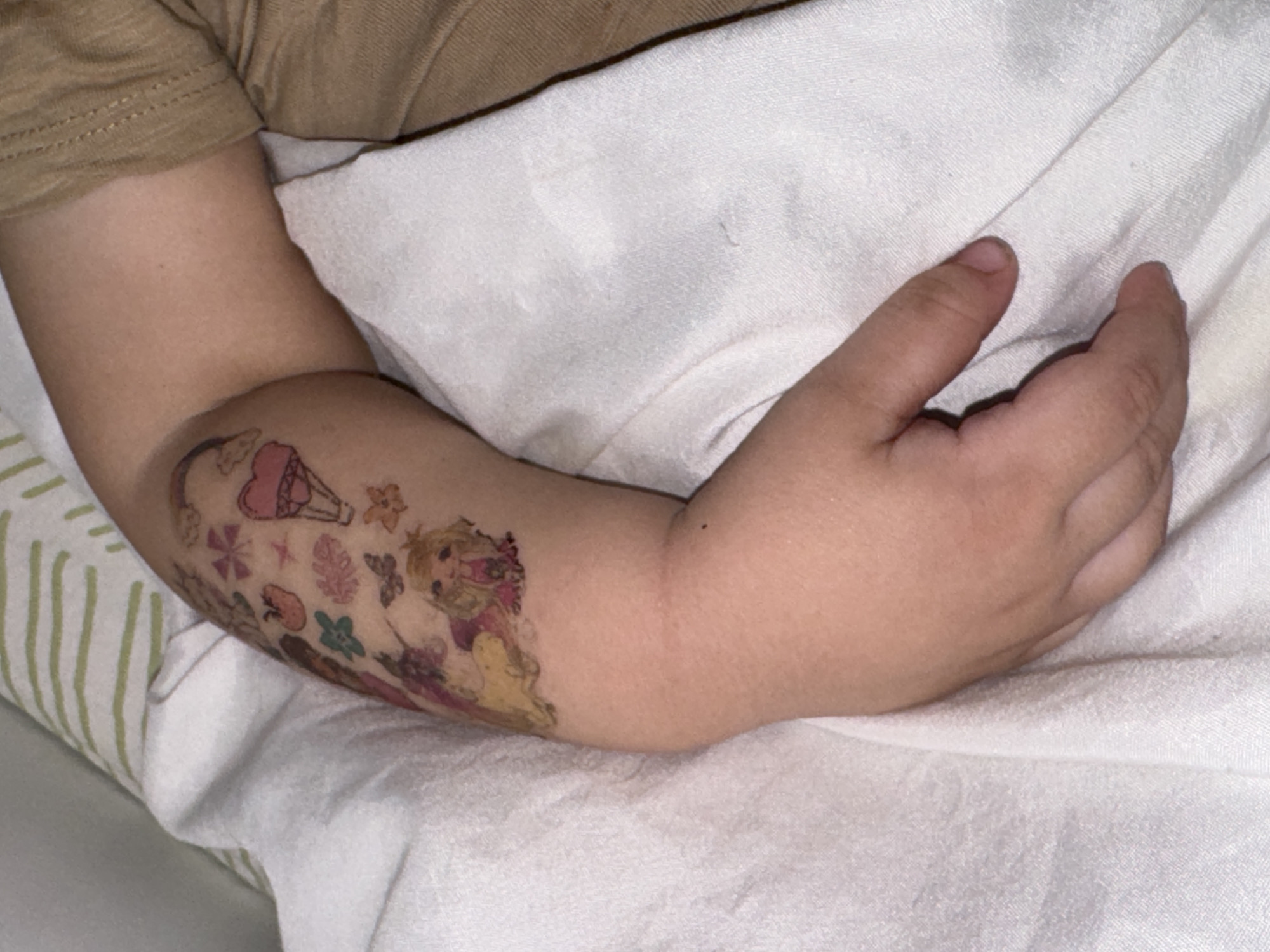 A close-up shot shows a child's left arm resting on a white surface, likely a bed or blanket. A colorful temporary tattoo adorns the arm, featuring images of a hot air balloon, flowers, and cartoon characters. The child's hand is positioned with fingers slightly curled. The context or location of the image within Chaikovsky, Russia, is not discernible from the visual content.