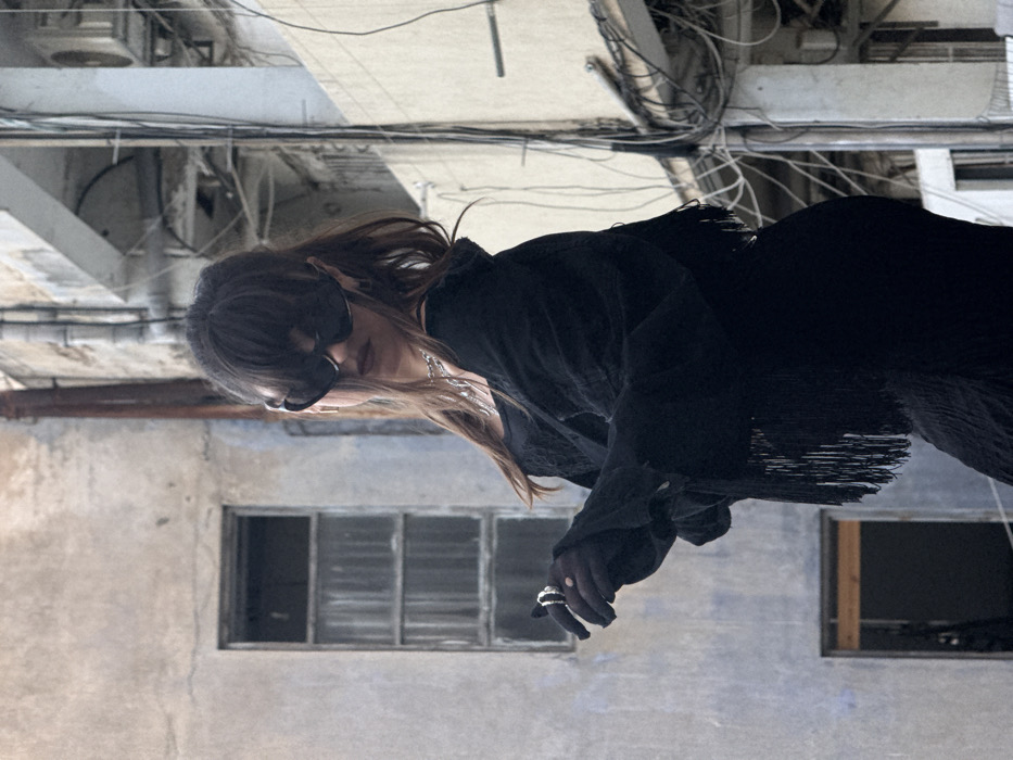 The image depicts a person in an urban environment within Tel-Aviv, Israel. The central subject, positioned slightly to the right, is a person with dark hair styled with bangs, wearing large, dark, cat-eye sunglasses that obscure the eyes and upper face. The individual is dressed in an entirely black ensemble, consisting of a textured undershirt, a loose-fitting jacket or cardigan with long fringes along its lower edge, and a textured black lower garment. A silver-colored chain necklace is visible around the neck. The subject's gloved left hand, raised slightly, displays a prominent multi-band ring featuring dark and light elements. The person maintains a posed posture, looking towards the viewer.
The background comprises two distinct building facades. To the left, a weathered, light-grey or blue-grey stucco wall displays signs of age and discoloration. It features a tall, narrow window with multiple vertical panes, its glass appearing opaque or reflective, and a partially visible dark opening below it. A vertical pipe, possibly rusted, runs down this wall. To the right, a lighter-colored concrete structure is extensively covered by a dense network of black electrical wires, cables, and conduits, illustrating exposed utility infrastructure common in older city districts. The scene conveys a utilitarian and somewhat aged urban aesthetic.
