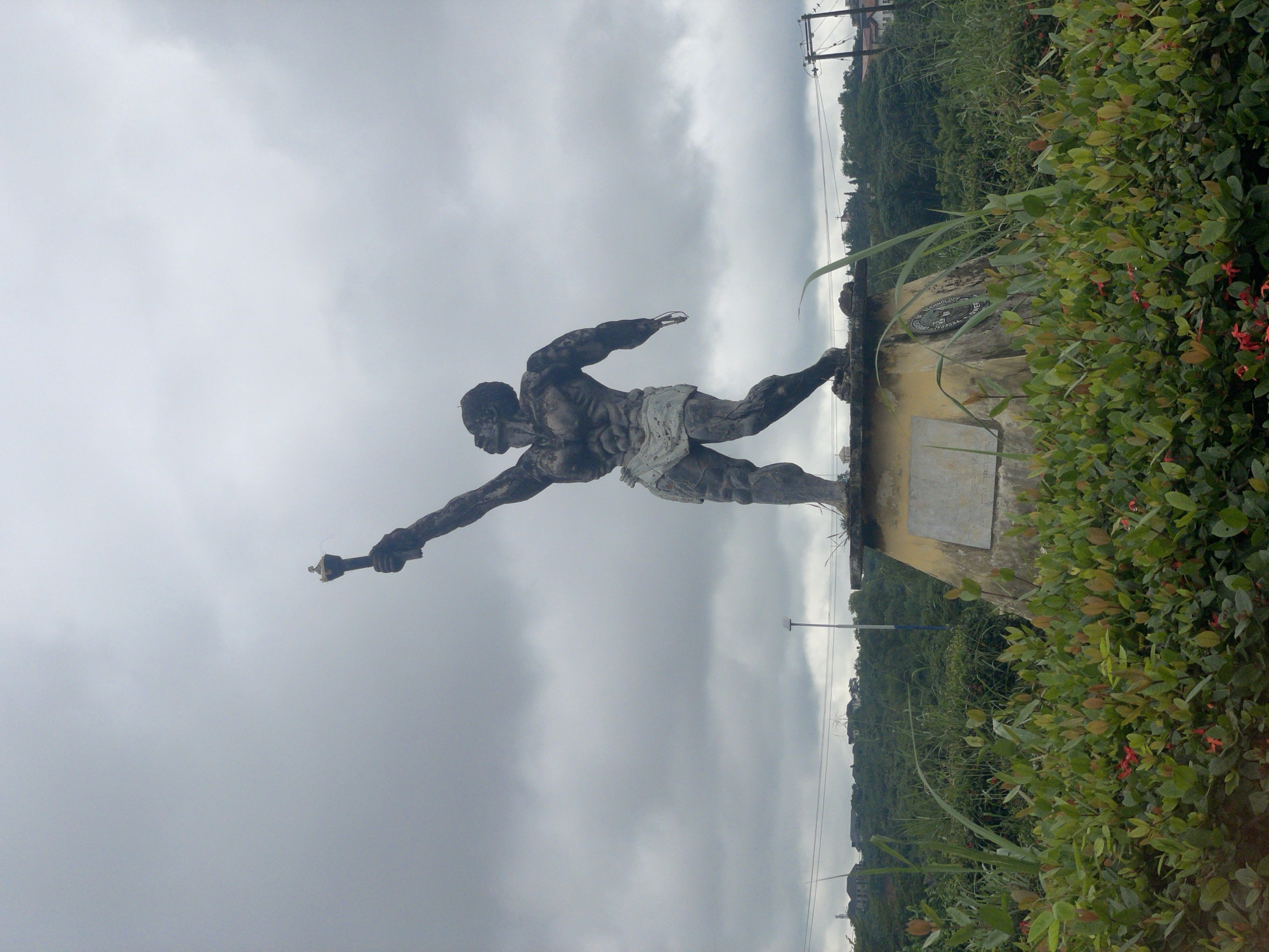 In Ihiagwa, Nigeria, a dark, weathered, full-body statue of a muscular man stands prominently against a cloudy sky. The man is depicted with a loincloth and holding a torch aloft in his right hand, its tip appearing to have a small crown or finial. The statue rests on a multi-tiered, weathered pedestal, which has a plaque on its front. The statue and pedestal are surrounded by lush green vegetation, including bushes with small red flowers in the foreground and taller grasses and trees in the background. In the distance, several buildings are visible on a green, hilly landscape, along with power lines and a street lamp. The overall atmosphere is somber due to the overcast sky and the weathered appearance of the statue.