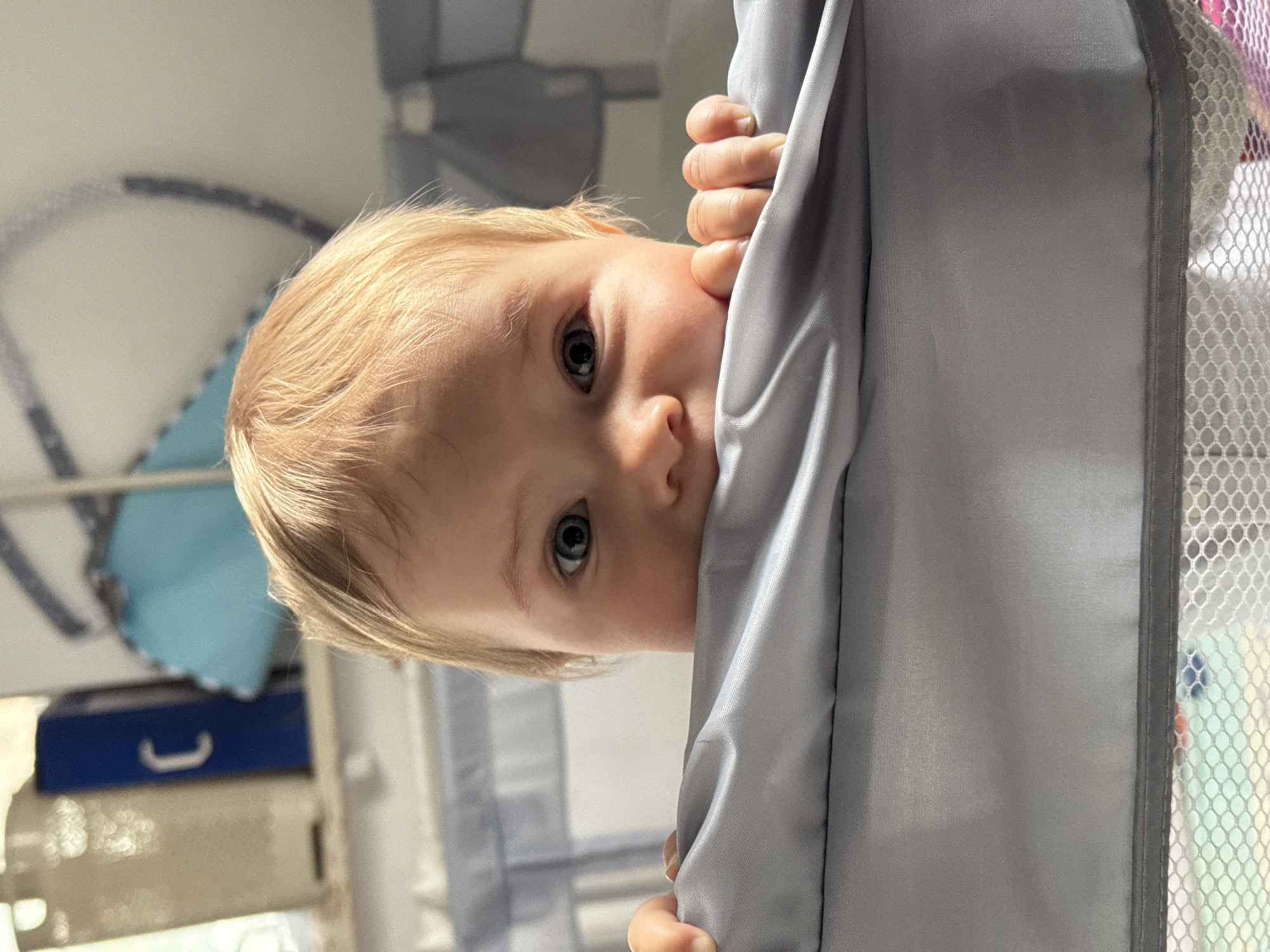A close-up, low-angle shot shows a baby peeking out from behind a gray fabric, likely a crib or playpen. The baby has fair skin and light blonde hair. Only the left side of the baby's face is visible, with a large blue eye looking directly at the camera. The baby's left hand is gripping the fabric. The background is softly blurred, showing what appears to be nursery items. A blue bin with a white handle is visible on a shelf to the left, and a pale blue object with a scalloped edge is behind it. The lighting suggests it is daytime. The overall scene evokes a sense of curiosity and playfulness.