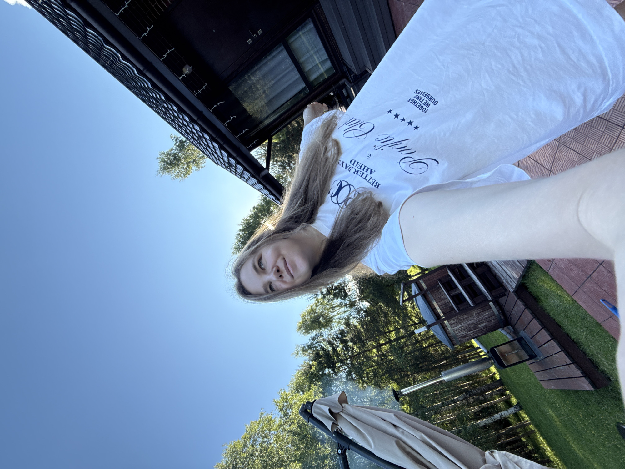 The image depicts a female subject in an outdoor setting identified as Копнино, Russia, captured from a low, tilted self-portrait perspective on a clear, sunny day. The subject, a young woman with long, light brown hair, is positioned in the lower right and center of the frame, looking upwards and to the right with a slight smile. She wears a loose-fitting white t-shirt featuring dark blue text: "BETTER DAYS AHEAD" with a circular logo above it, "PACIFIC Club" with five stars below, and "TOGETHER WE FIND OURSELVES" in smaller print.
The background reveals a verdant environment with numerous trees, including birches and conifers, under a vast, cloudless blue sky. To the left, a large, closed beige outdoor umbrella stands next to a metal chimney stack connected to an unseen heating unit, situated on wooden decking. Further right, a wooden hot tub with external steps is visible, also on wooden decking. In the upper right and center, a dark brown house or cabin with a black, tiled roof is present, featuring a window and what appear to be decorative string lights hanging along the eaves. The ground consists of green grass, sections of reddish-brown patterned paving, and wooden decking. Sunlight casts bright illumination across the scene, indicating daytime.