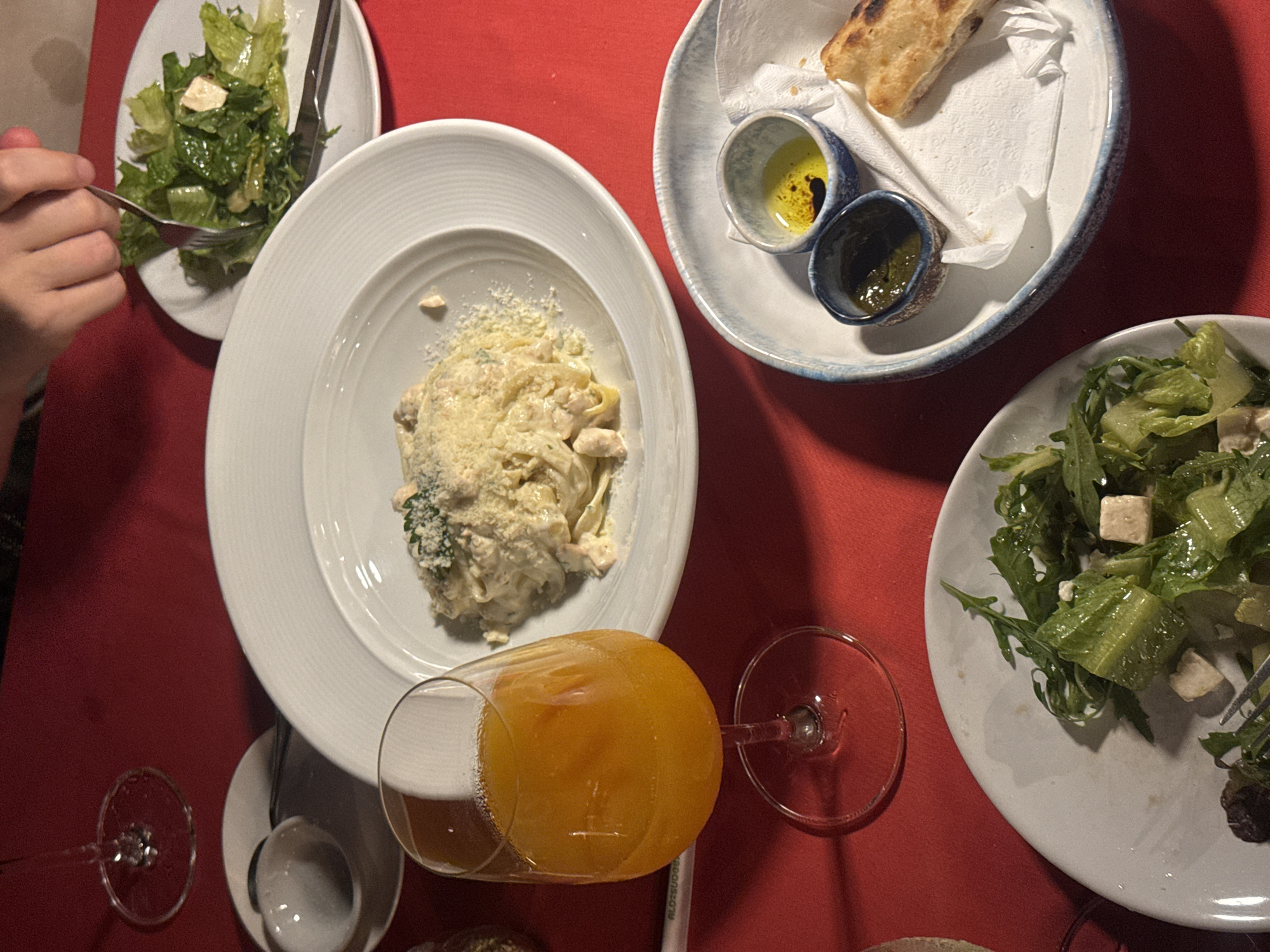 This overhead, dimly lit photo depicts a table setting with several plates of food and drinks, suggesting a meal is in progress. The scene is set against a vibrant red tablecloth.
On the left, a hand is visible reaching for a forkful of salad from a white plate. The salad appears to be a light green variety, possibly romaine lettuce, with white cubes that could be cheese. Beside this plate is another white plate with a pasta dish, likely fettuccine or a similar flat noodle, covered in a creamy sauce and grated cheese.
To the right of the pasta, a shallow, blue-rimmed bowl contains two small dipping cups. One cup holds a yellowish liquid, likely olive oil with balsamic vinegar, and the other contains a darker green sauce. A piece of toasted bread is resting on a white napkin within the bowl.
Further to the right, another white plate holds a salad with mixed greens, including what looks like arugula, and more of the white cheese cubes.
In the foreground, a stemmed glass is filled with a bright orange liquid, possibly juice or a cocktail, with a foamy head. Another empty stemmed glass is visible to the left of the drink. Two white bowls, one containing a spoon, are also present on the table.
The overall impression is that of a casual dining experience. The lighting and the presence of multiple plates suggest this is likely an indoor setting, possibly a restaurant, during the evening. There is no visible text in the image.