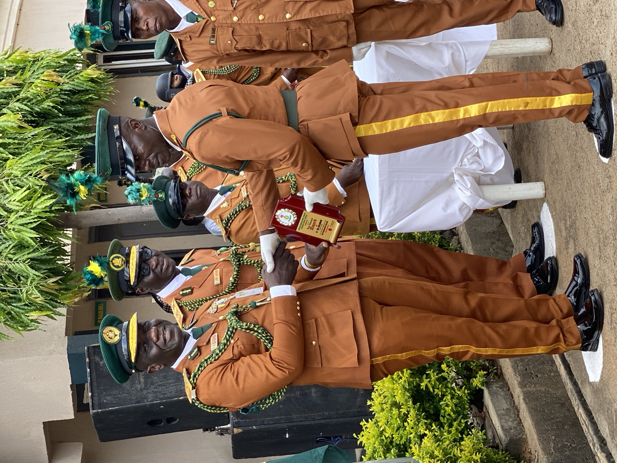The image depicts an award ceremony taking place outdoors in Kaduna, Nigeria. The central focus is on two men in ceremonial military uniforms. The man on the left, wearing a tan uniform with green trim and a green cap adorned with a gold emblem, is smiling and extending his hand to shake the hand of the man on the right. The man on the right, also in a similar tan uniform with green trim and a green cap with a gold emblem and green and yellow plumage, is holding a red award plaque with gold trim and a green emblem. He is wearing white gloves. Several other men in similar uniforms are visible in the background, some with their faces partially obscured. They are standing in front of a building with a light-colored facade and a few large black speakers. Green foliage and trees are present in the background and to the left of the frame. A white tablecloth covers a surface in the middle ground, likely a table. The ground appears to be paved, with some concrete elements. The overall scene suggests a formal event, possibly a graduation or recognition ceremony.