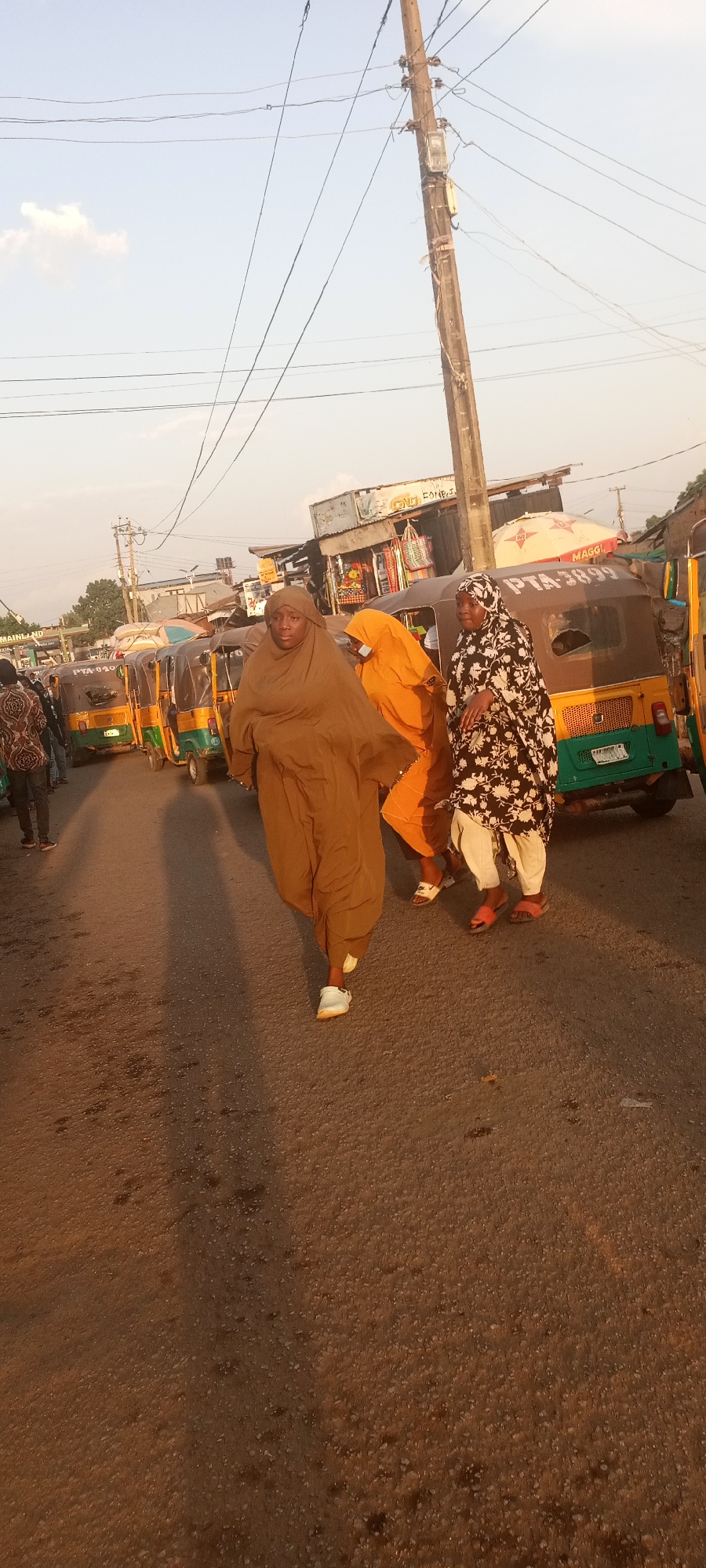 The image shows an urban street scene in Jos, Nigeria. Three women are walking on the street: one is in a brown hijab and white sneakers, another in an orange outfit, and the third is wearing a black and white floral dress and sandals. Behind them, several yellow and green auto-rickshaws (tuk-tuks) are parked. There is a wooden utility pole in the background. Also visible are storefronts in the distance. The street surface is asphalt.