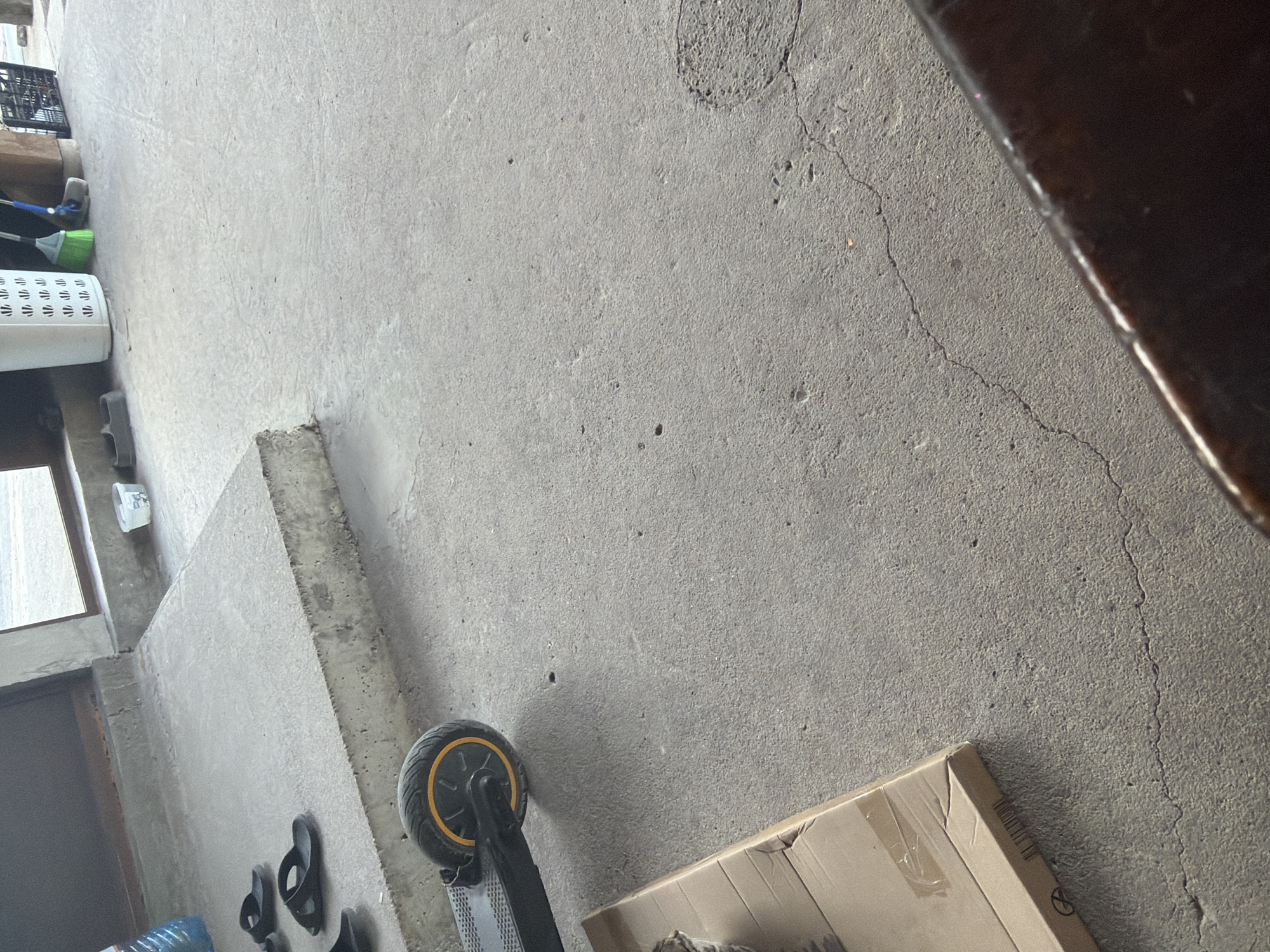 The image depicts a concrete floor and wall, possibly in a garage or storage area, as inferred from the lack of finished surfaces and presence of stored items. The floor has cracks and some discoloration. A black and yellow-wheeled scooter is visible near the bottom, along with a cardboard box. Other objects include sandals, plastic bottles, a white plastic container, and cleaning supplies. The location cannot be determined from the provided information. There are no people visible in the image.