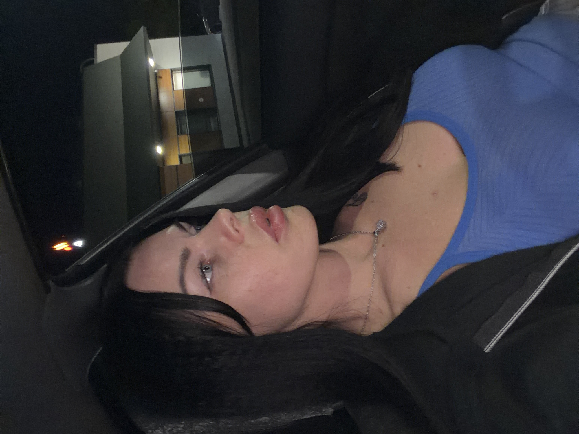 The media file is a close-up, horizontally oriented photograph featuring a young woman lying down, looking upwards and to the left. She has long, dark hair that frames her face and shoulder. She is wearing a bright blue top and a silver necklace with a small pendant. Her lips are slightly parted and appear to be glossy. Her eyes are open, and her gaze is directed out of frame.
The setting appears to be inside a vehicle at night, suggested by the dark interior and the illuminated building visible through a window in the background. The building has modern architectural elements with several lit windows and a distinctive angled facade. There are some glowing orange lights, possibly from another vehicle or streetlights, visible on the left side of the frame. The overall lighting is dim, with the primary light source seemingly coming from the building and possibly a flash used for the photograph, which highlights the woman's face and upper body.
The woman's expression is difficult to definitively interpret due to the angle and lighting, but her slightly open mouth and upward gaze could suggest a moment of reflection, surprise, or mild distress. The image seems to capture a candid moment, possibly during a car ride or a pause in activity. There is no visible text in the image. The location context provided is Ufa, Russia.