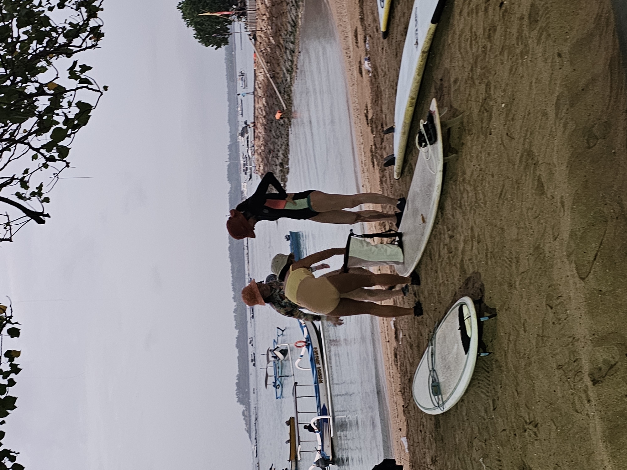 The image depicts a cloudy day at a beach in Denpasar, Indonesia, likely near Sanur. In the foreground, three individuals are on a sandy beach, with two surfboards laid out on the sand. Two of the individuals are wearing swimsuits and hats, appearing to be preparing for or finishing a surfing session. One person is wearing a black and colored wetsuit. In the background, the ocean is calm, dotted with numerous traditional Indonesian fishing boats, some of which are docked at a rocky pier. The sky is overcast, suggesting either early morning or an approaching storm. The scene conveys a relaxed, watersport-oriented atmosphere typical of coastal areas in Bali.