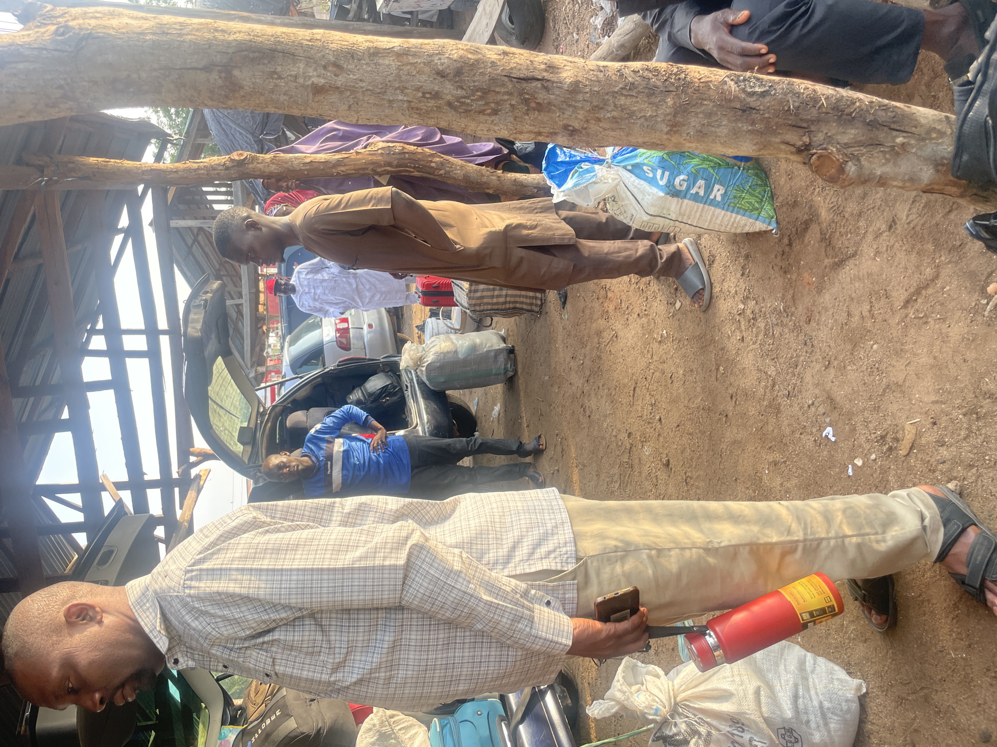 The image depicts a dusty outdoor area, possibly a marketplace or transportation hub, in Wauro Jabbe, Nigeria. The scene is characterized by the presence of several individuals, luggage, and vehicles. Sunlight filters through a rustic wooden shelter overhead, casting long shadows.
In the foreground, a man wearing a checkered shirt and light-colored trousers stands facing left, holding a red water bottle in his left hand and a mobile phone in his right. To his right, another man in a blue long-sleeved shirt and dark trousers stands with his hands on his hips, looking towards the left. Behind him, the open trunk of a car reveals luggage.
Further into the background, a group of people are gathered. One man wears a white traditional outfit and a red cap, while another person in the foreground wears a brown traditional outfit. Several bags and suitcases are visible, including a large white bag with blue lettering that appears to say "SUGAR". The ground is covered in dirt and dust, with some scattered debris. The overall impression is one of activity and preparation, likely related to travel or commerce.