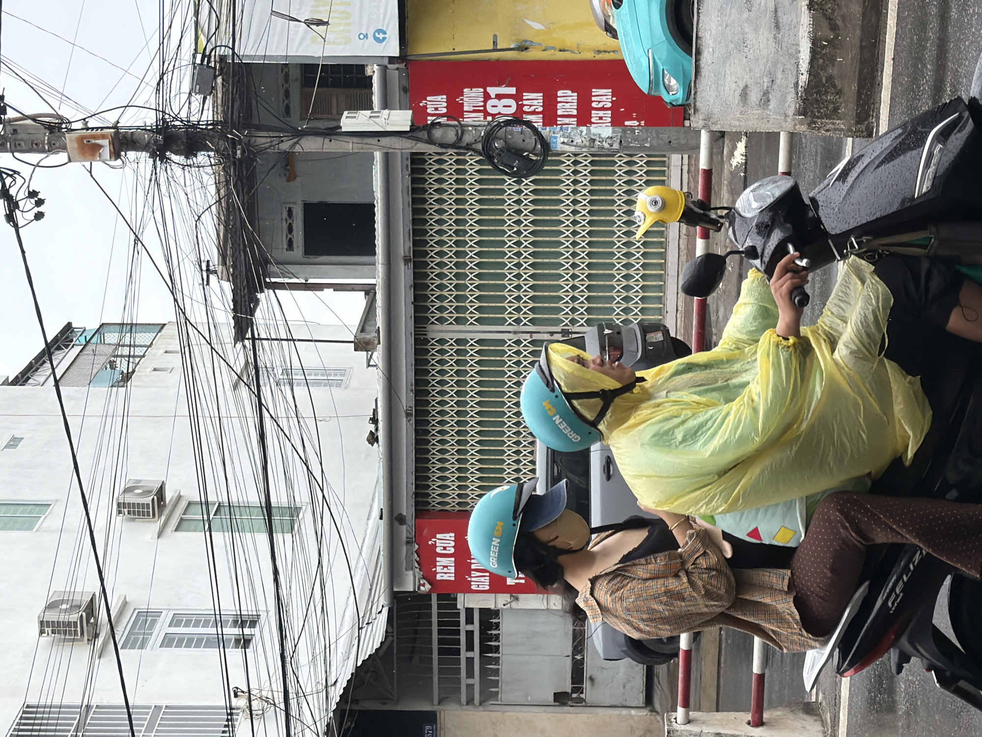 In Nha Trang, Vietnam, two individuals, likely a driver and a passenger, are riding on a dark-colored scooter. The driver, wearing a bright yellow hooded raincoat and a teal helmet with "GREEN SM" branding, is operating the scooter. The passenger, positioned behind the driver, is wearing a plaid shirt over a dark top, brown pants with a polka-dot pattern, and a teal helmet also bearing the "GREEN SM" logo, with a tan face mask and a blue baseball cap visible beneath the helmet. A light blue bag with colorful geometric shapes is held by the passenger.
The scooter is equipped with a bright yellow, duck-shaped accessory attached to the front. The scene unfolds on a wet, asphalt road, indicating recent or ongoing rain. The background features a multi-story building with white walls and windows, alongside a building with a red facade displaying Vietnamese text and a metallic roll-up door. Overhead, a dense network of electrical wires crisscrosses the sky, attached to a utility pole. Further down the street, a turquoise-colored vehicle is partially visible. The overall environment suggests a typical urban street scene in Vietnam.
