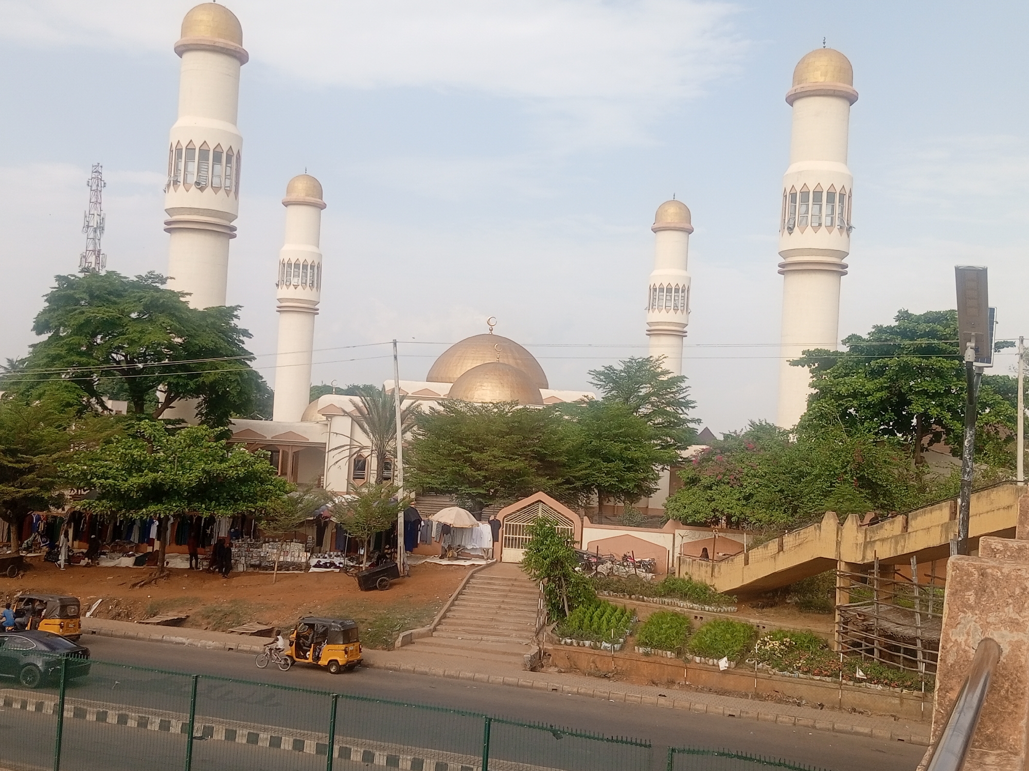 The image shows a mosque with four tall, white minarets capped with gold domes. A larger gold dome tops the main structure of the mosque. The mosque is set back from a road, with trees and some market stalls in front. A pedestrian overpass is visible on the right side of the frame. Three-wheeled vehicles are on the road. The sky is overcast.