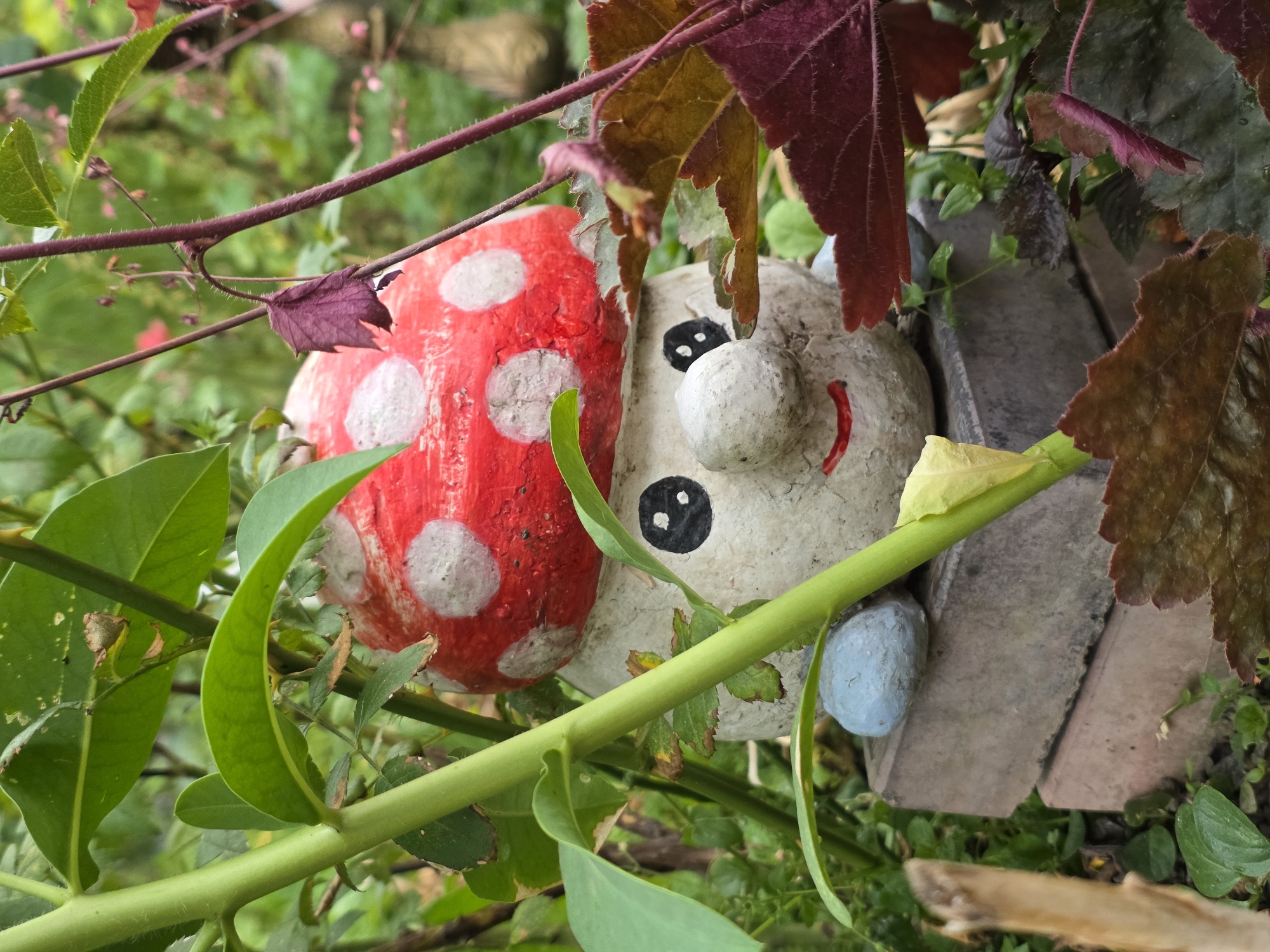 An eye-level, medium close-up shot shows a whimsical garden gnome with a red, white-spotted mushroom cap head. The gnome's round face is painted with black eyes and a red, smiling mouth. It is perched on two grey bricks, with a light blue stone resting on the top brick to its left. Lush green foliage and dark reddish-purple leaves surround the gnome, partially obscuring it. The background is blurred, suggesting a garden setting in an unknown location.