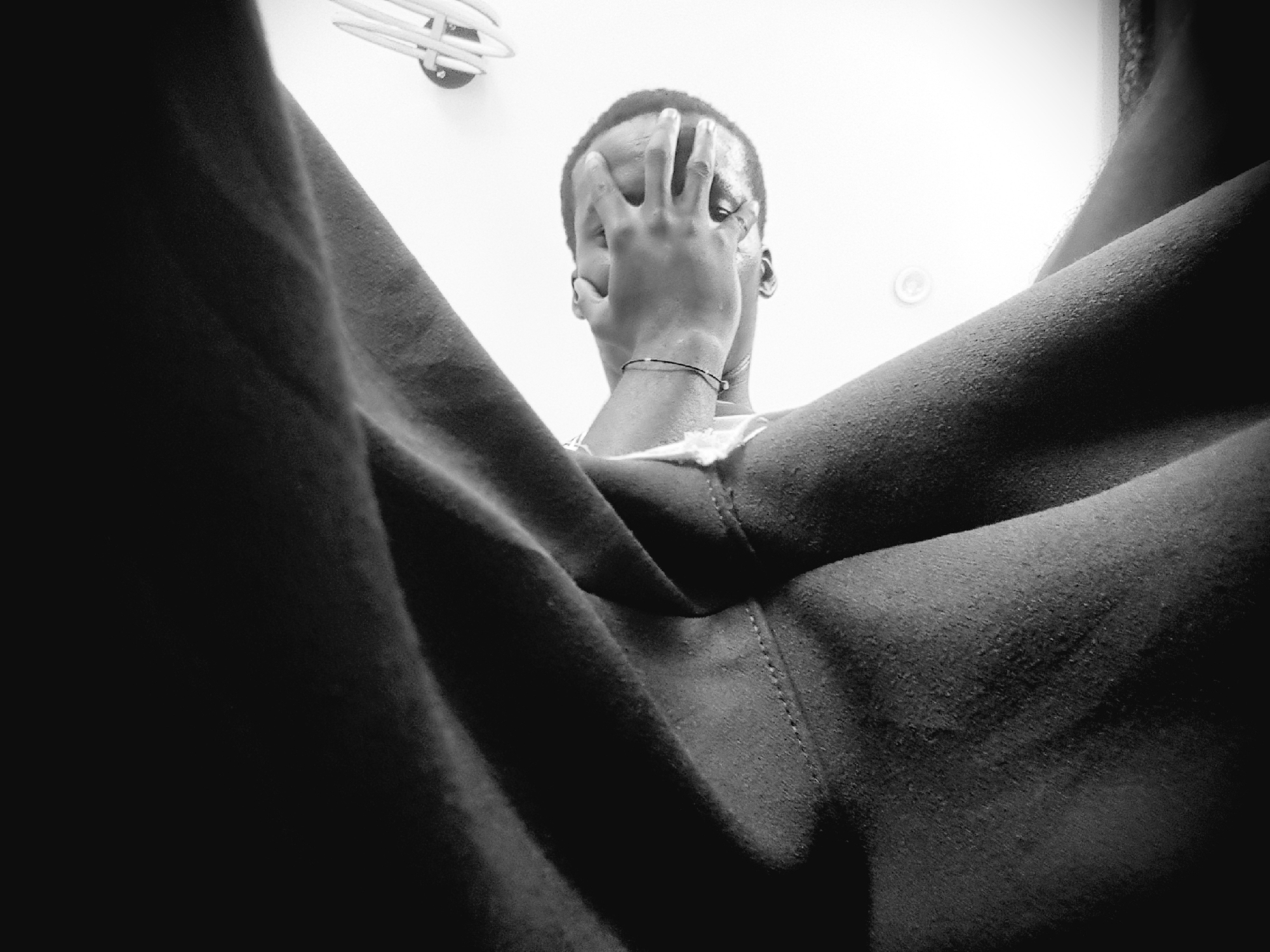 The photograph is a black and white, low-angle shot of a person's head and shoulders, seen from below and tilted to the side. The person, who appears to be an adult male with dark skin, is covering his face with his right hand. He is wearing a thin bracelet on his left wrist. The subject is positioned against a brightly lit background, possibly a wall or ceiling, with a ceiling fixture visible in the upper right corner. The left side of the image is dominated by a dark, textured fabric, likely clothing, that drapes over the person's head and shoulders, creating a sense of partial concealment. The lighting creates strong contrasts and shadows, emphasizing the textures of the fabric and the contours of the person's face and arm. The image appears to be taken indoors. There is no discernible information about the location in the city of Narayi, Nigeria within the visual content of the image.