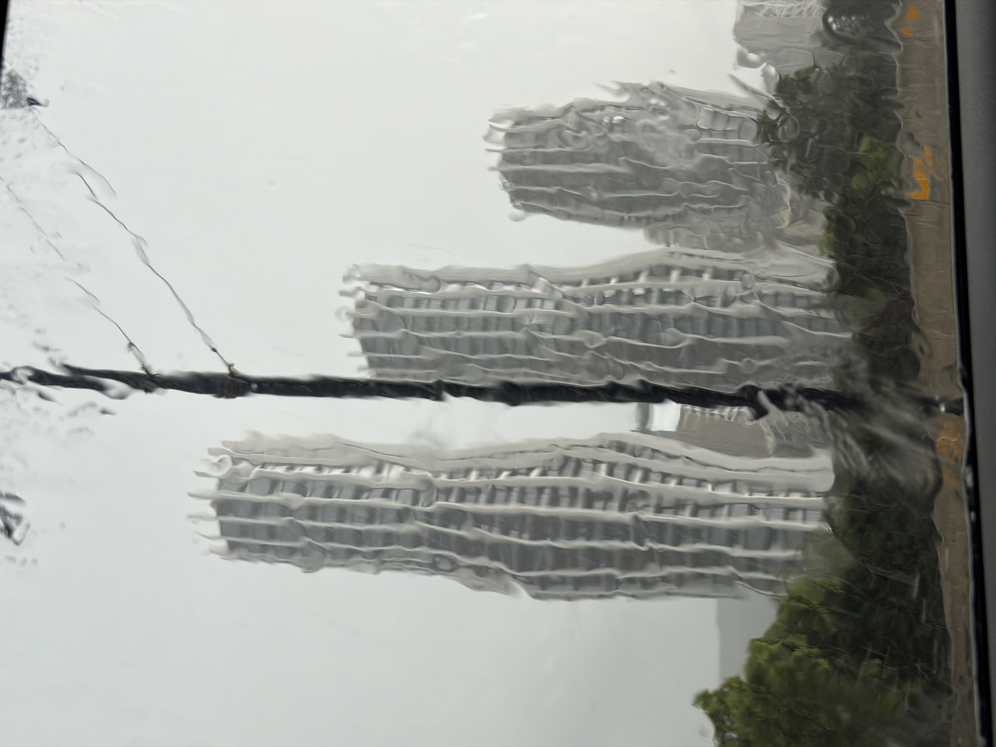 The image depicts a view through a rain-streaked window. Outside the window, partially obscured by the rain, are several tall, light-colored buildings with a grid-like pattern, suggesting offices or apartments. The buildings are next to trees. A dark wire stretches across the foreground of the window. The scene is blurry, likely due to the rain and focus. The environment suggests an urban setting, specifically Tbilisi, Georgia.