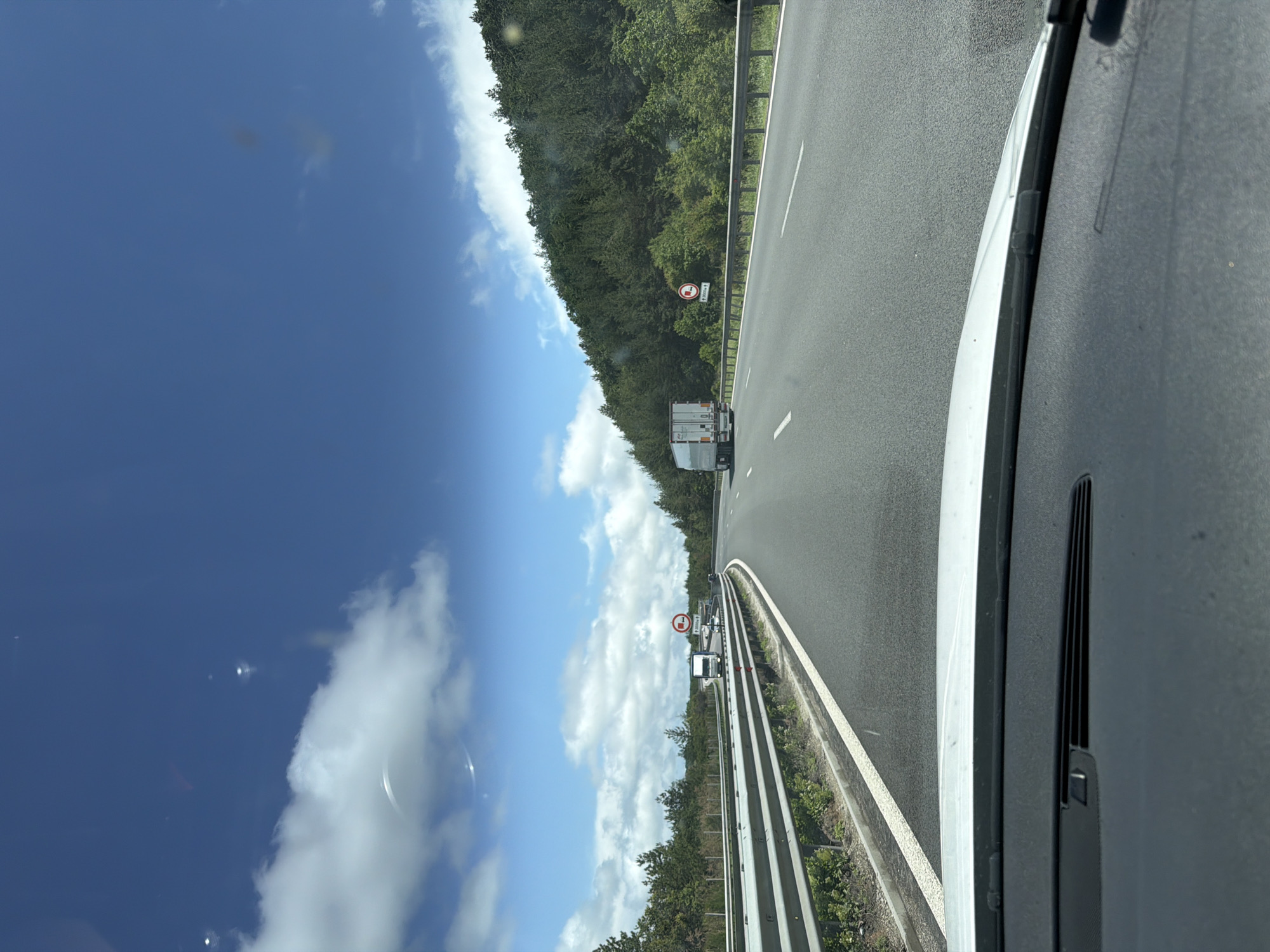The image captures a daytime scene from the interior of a vehicle traveling on a highway. The perspective is from the front passenger or driver's side, looking forward and slightly to the right, revealing a portion of the car's white A-pillar and black dashboard/windshield area on the right.
The scene features a multi-lane asphalt highway stretching into the distance under a bright blue sky dotted with scattered white cumulus clouds. A dense green forest or treeline runs along the right side of the highway, separated by a metal guardrail.
In the distance, a white semi-trailer truck is visible in the right lane. Further ahead, another smaller vehicle can be seen. A prominent red-bordered circular road sign is posted on the right side of the highway, displaying a truck symbol and the text "3.5 t", indicating a weight restriction (likely "no entry for vehicles over 3.5 tons" or similar). Another, less discernible, red-bordered circular sign appears further down the road.
The weather is clear and sunny, suggesting a pleasant day for travel. The location context is Krushovitsa, Bulgaria.