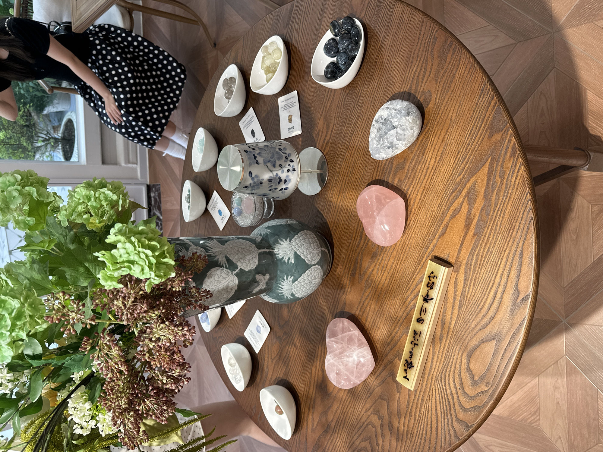 An overhead shot reveals a round, wooden table displaying an array of items, including polished stones and decorative objects. Several small, white, elliptical bowls contain different types of stones. There are also two cups, one decorated with white and blue floral patterns, and the other transparent with visible liquid inside. Adjacent to these are identification cards with Chinese characters. Near the edge of the table are three pink, heart-shaped stones. A wooden block with additional Chinese writing rests on the table. A large vase filled with green and brown floral arrangements is positioned near the edge of the table. In the background, a person wearing a black and white polka-dot dress can be partially seen. The scene is set indoors, with a parquet floor visible in the background. The location of the photo is the city of Liangjiang, China.