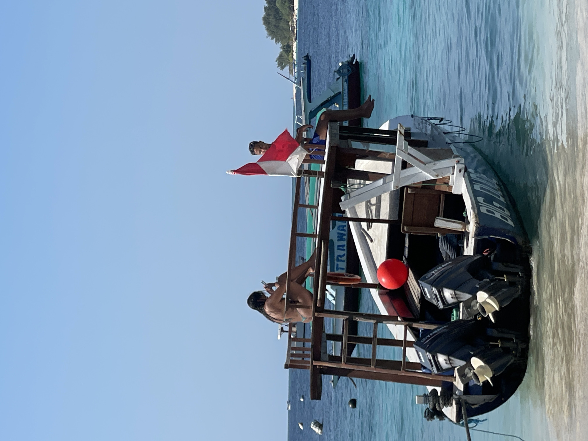 Two individuals are seated on a wooden platform extending from a boat on the water. The person on the left, a woman with dark hair, is wearing a bikini and looking at her phone. The person on the right, a man wearing blue shorts, is holding an Indonesian flag. The boat has two outboard motors and the name "BIG DUD" is visible on its hull. The water is clear and turquoise, and other boats and a tropical island with trees are visible in the background under a clear blue sky.