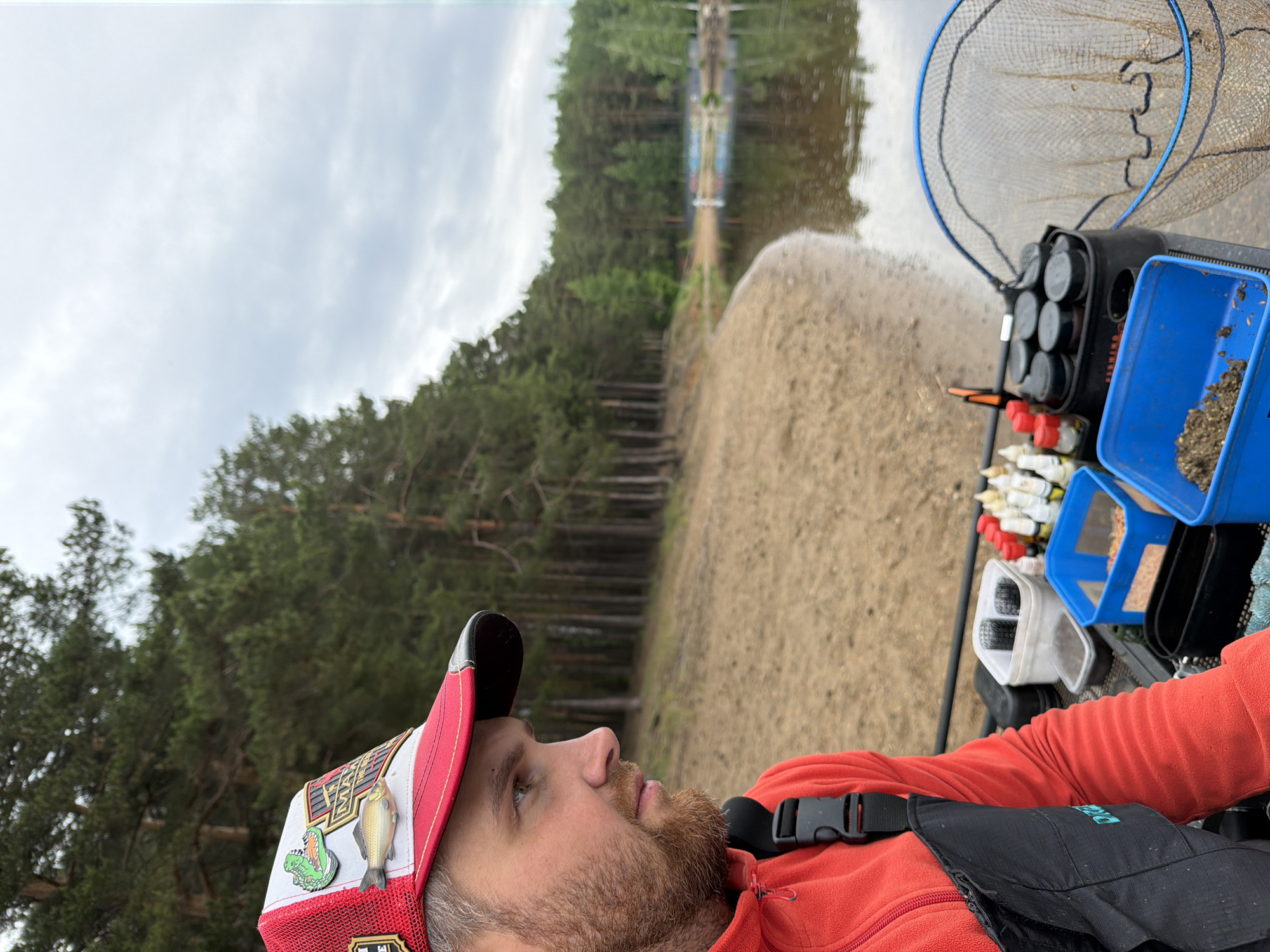 The image shows a man, likely a fisherman, wearing a red and white cap adorned with pins of a fish and an alligator. He has a beard and is wearing a red fleece jacket and a black vest. The background reveals a shoreline with trees and water, suggesting a river or lake. Fishing equipment, including a net and tackle boxes, is visible next to the man. The location is Chaikovsky, Russia.
