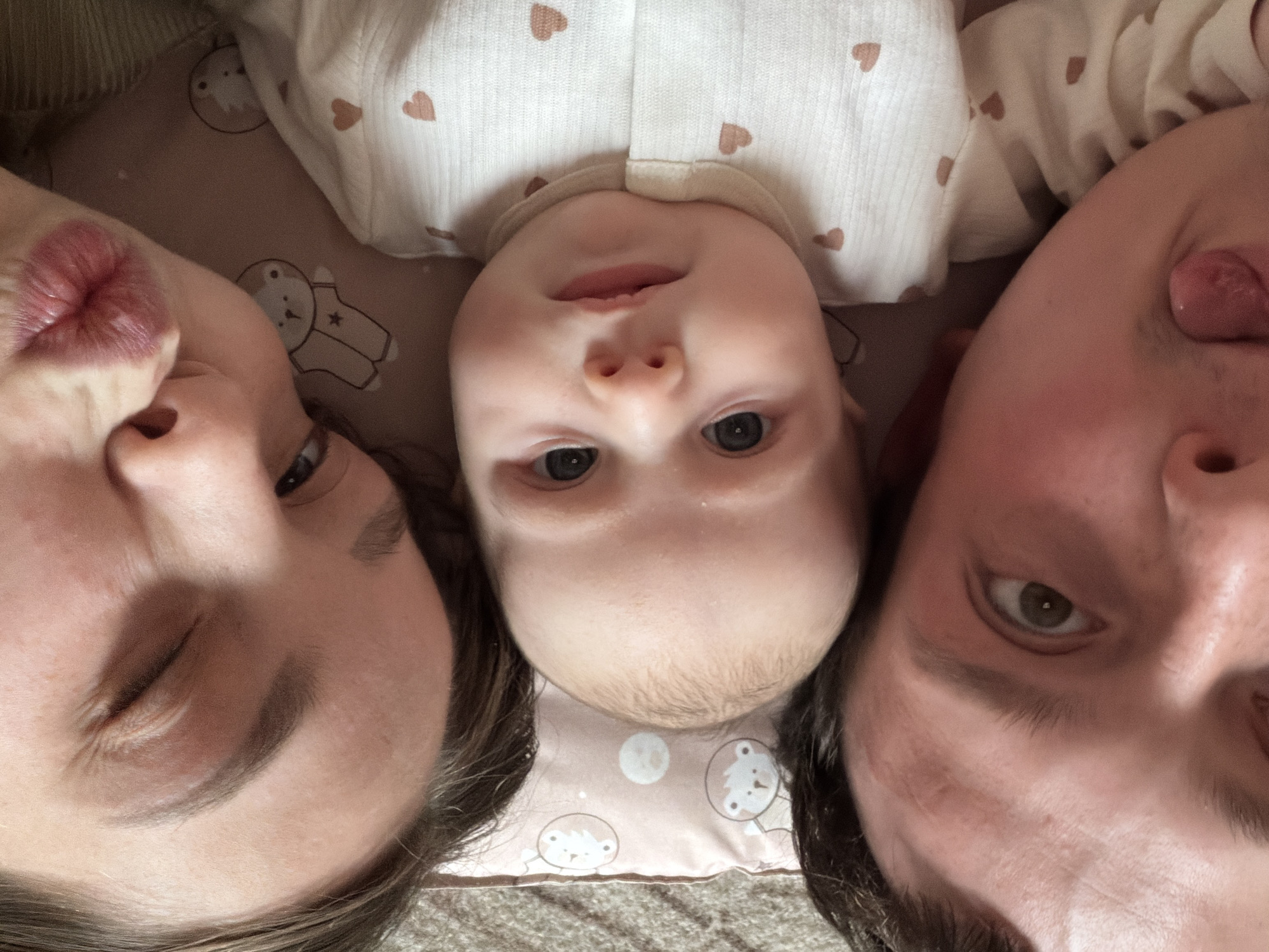A close-up, top-down photograph depicts a baby lying between two adults. The baby is centered, with a white outfit patterned with small brown hearts. The woman on the left is puckering her lips towards the camera, with one eye closed. The man on the right is sticking out his tongue. All three subjects appear to be lying on a light pink blanket or sheet with a pattern of cartoon bears. The background appears to be a simple interior. There is no indication of the city Chaikovsky, Russia.
