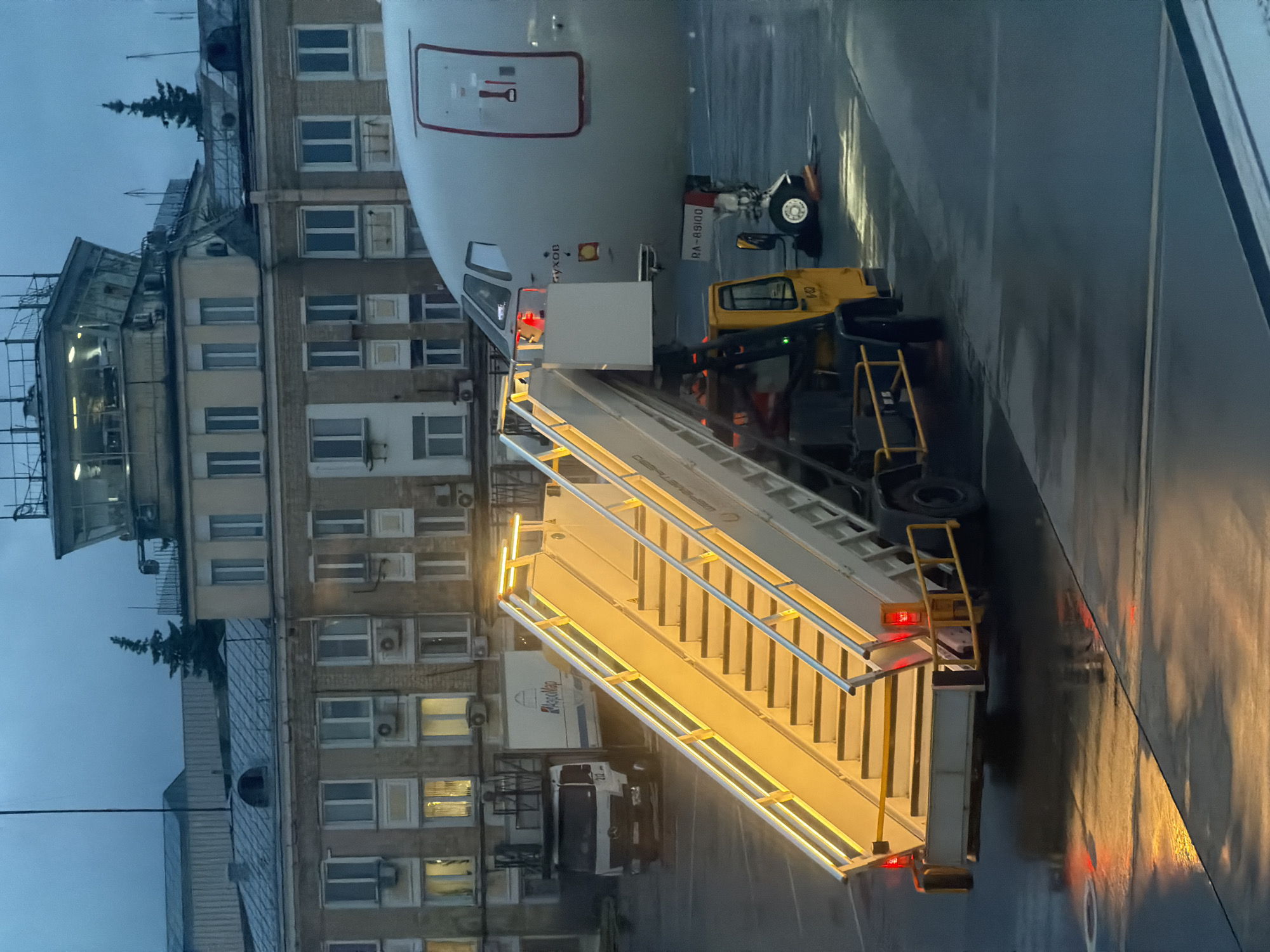 This image, taken at Sheremetyevo International Airport in Khimki, Russia, depicts an airplane parked at an airport gate during what appears to be dawn or dusk. The sky is overcast and a muted blue-grey. In the foreground, a mobile passenger staircase with bright yellow lights runs diagonally from the bottom left towards the center right, leading up to an airplane. The staircase is attached to a vehicle. To the left of the staircase, a white truck with a logo is visible, and behind it, a light-colored airport building with numerous windows and air conditioning units stretches across the background. Above the building, a control tower with multiple windows and antennas is prominent. The ground is wet, reflecting the lights from the staircase and the aircraft. The airplane in the center right is white with a red stripe and appears to be a Tupolev Tu-204, as indicated by the visible "ТУ-204" marking and the registration number "RA-64100" on the fuselage. The scene is devoid of visible people, suggesting the airport is either just beginning operations or winding down for the day.