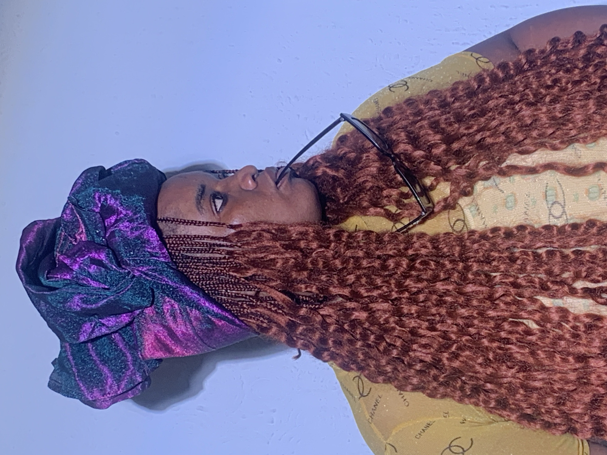 A person with dark skin is depicted in a portrait-style image, oriented sideways so their head points upwards. They are wearing a shimmering purple and green iridescent headwrap or turban. Long, voluminous reddish-brown hair, styled in braids or twists with a curly texture, extends across the frame. Black-framed sunglasses are perched on their face, with the arms resting against their mouth. The individual is dressed in a light yellow or gold top, prominently featuring a repeating pattern of the "CHANEL" text and the brand's interlocking 'CC' logo. Their eyes are open and directed upwards, conveying a thoughtful or composed expression as if posing for the camera. The background is a plain, light blue wall, suggesting an indoor setting. The image offers no specific time of day or weather cues, though the subject's attire and appearance align with the provided location context of Barakin Akawo, Nigeria.