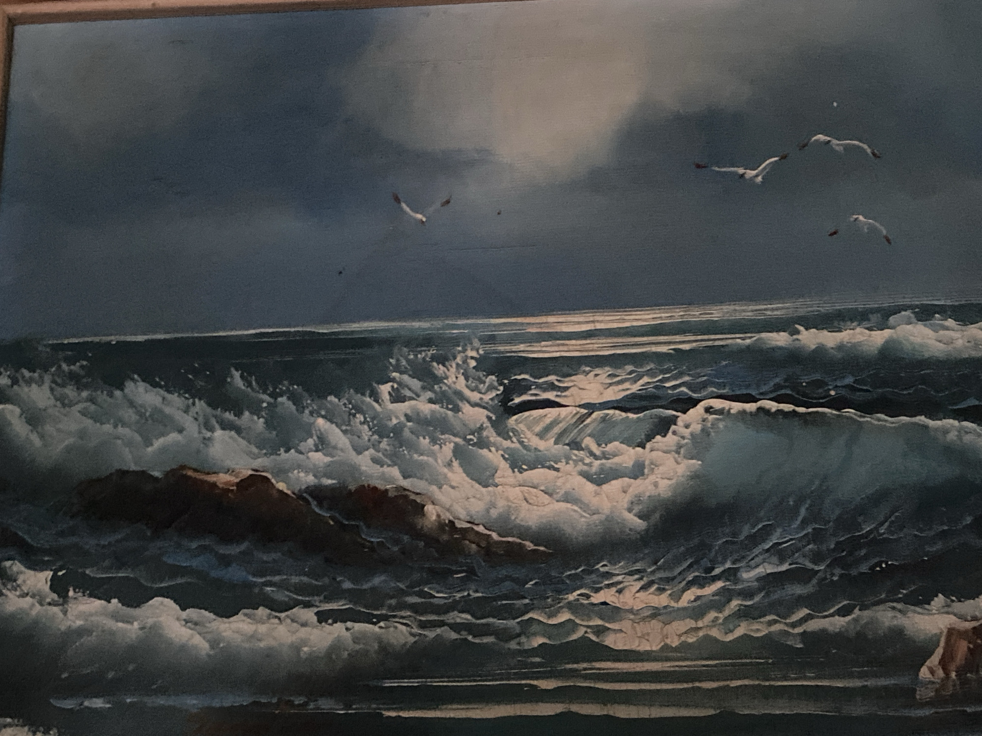 The image is a painting depicting a stormy seascape. Turbulent waves crash against a rocky outcropping in the foreground. Above, the sky is filled with dark, ominous clouds. Several seagulls are in flight against this dramatic backdrop. The painting evokes a sense of raw, natural power and untamed beauty. There is no indication of Tinaquillo, Venezuela in the image.
