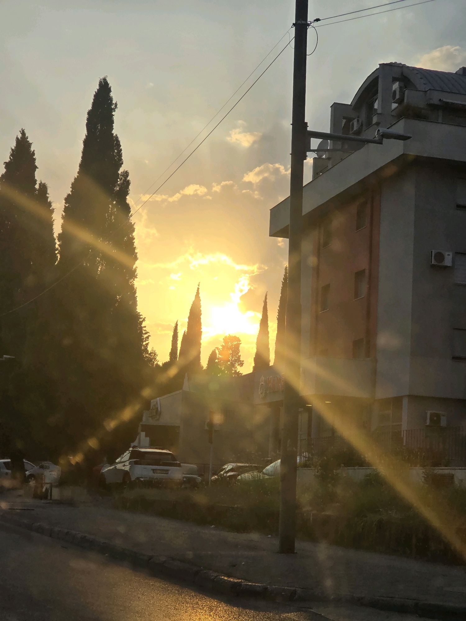 The image shows a street scene in Podgorica, Montenegro, during what appears to be sunset or sunrise. Tall, slender trees, possibly cypress, dominate the left side of the frame, silhouetted against a bright sky illuminated by the sun. The sun's rays cut diagonally across the scene, creating dramatic light shafts.
In the background, a modern building with a grey facade and a curved roof is visible on the right. Several windows and air conditioning units are apparent on its exterior. Adjacent to this building, another structure with a sign that is partially obscured but appears to have lettering on it, possibly a business or establishment.
On the street level, several cars are parked or driving. One white pickup truck is prominently featured in the middle ground. The street surface is wet, reflecting the light from the sky, suggesting recent rain. A utility pole with wires runs vertically through the center of the image. The overall impression is of an urban environment bathed in warm, low-angled light.