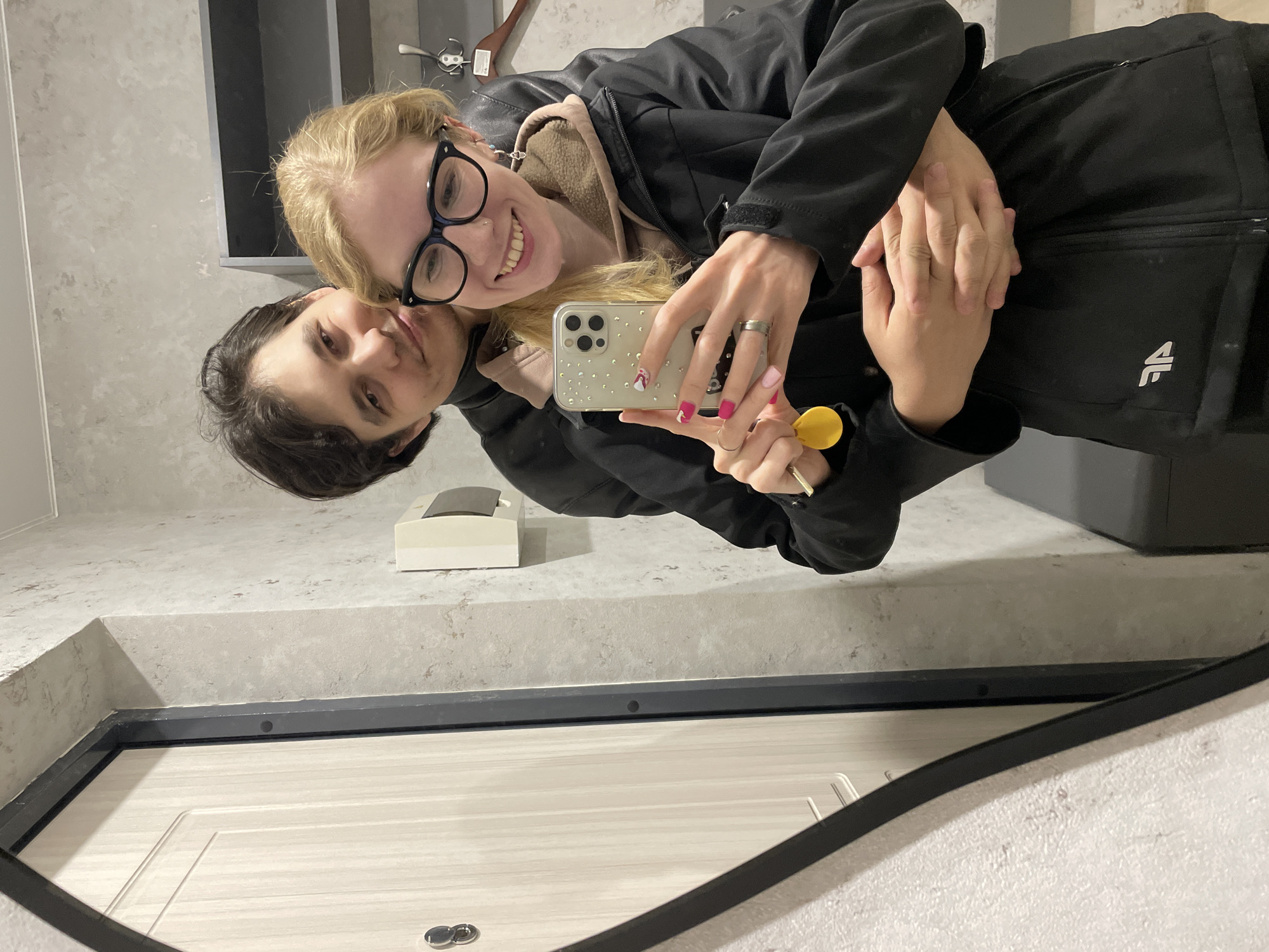 A couple takes an affectionate mirror selfie indoors, likely in Izhevsk, Russia. The image is rotated 90 degrees clockwise.
In the foreground, a blonde woman, wearing black-rimmed glasses, a black jacket over a beige hooded top, and bright pink and white nail polish, smiles broadly towards the camera. She holds a smartphone with a clear, sparkly case and a yellow key fob in her right hand. Her left arm is wrapped around the man beside her.
The man, with dark hair, rests his head against her and looks towards the camera with a subtle, gentle expression. He wears a black jacket with a white "AF" logo visible on the chest. Their hands are clasped together, suggesting intimacy.
The background, seen in the mirror's reflection, reveals a modern interior space. A gray, textured wall or countertop is visible behind them, along with a small white rectangular object and a darker cabinet or shelf. The lower part of the reflection shows a light-colored paneled door with a dark frame, and small metallic key rings on the bottom frame. The lighting suggests an indoor setting, likely daytime or well-lit by artificial lights. The overall mood is warm and joyful.