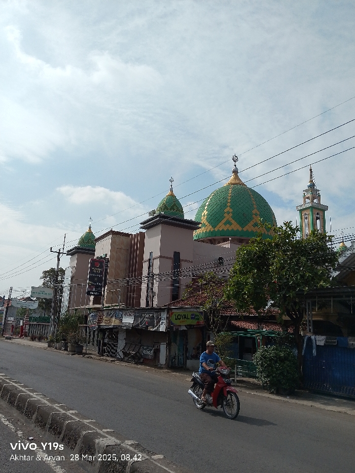 The image shows a street scene in Jatibarang, Indonesia (latitude: -6.476, longitude: 108.317). A man is driving a red motorcycle on a paved road. The road is lined with concrete curbs. There are buildings on both sides of the road, including a mosque with three green domes and a minaret. The mosque is partially obscured by a large tree with green leaves. There are power lines overhead and a cloudy sky above. There are signs on the building behind the tree that read "Loyal" and "Terang". Several small businesses can be seen next to the mosque. The image was taken on March 28, 2025, at 8:42 AM.