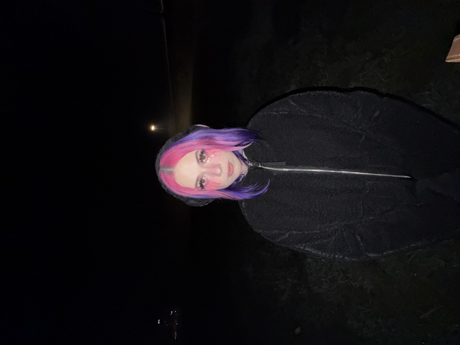 The scene is an outdoor, nighttime environment within the City of Zagreb, Croatia. A female individual is positioned centrally, facing the viewer directly with a neutral expression. She has shoulder-length hair, styled with a distinct pink center parting and purple side sections. Her makeup includes pink/red tear-like streaks embellished with glitter beneath each eye. She wears a dark, full-zip, high-collared jacket with a visible textured surface. The immediate foreground and surrounding area are dark and indistinct, suggesting unpaved or natural ground cover. In the distant background, a single bright light source is visible on the right, with several fainter, blurred lights appearing further to the left. No specific landmarks are visible to determine the precise location within the City of Zagreb.