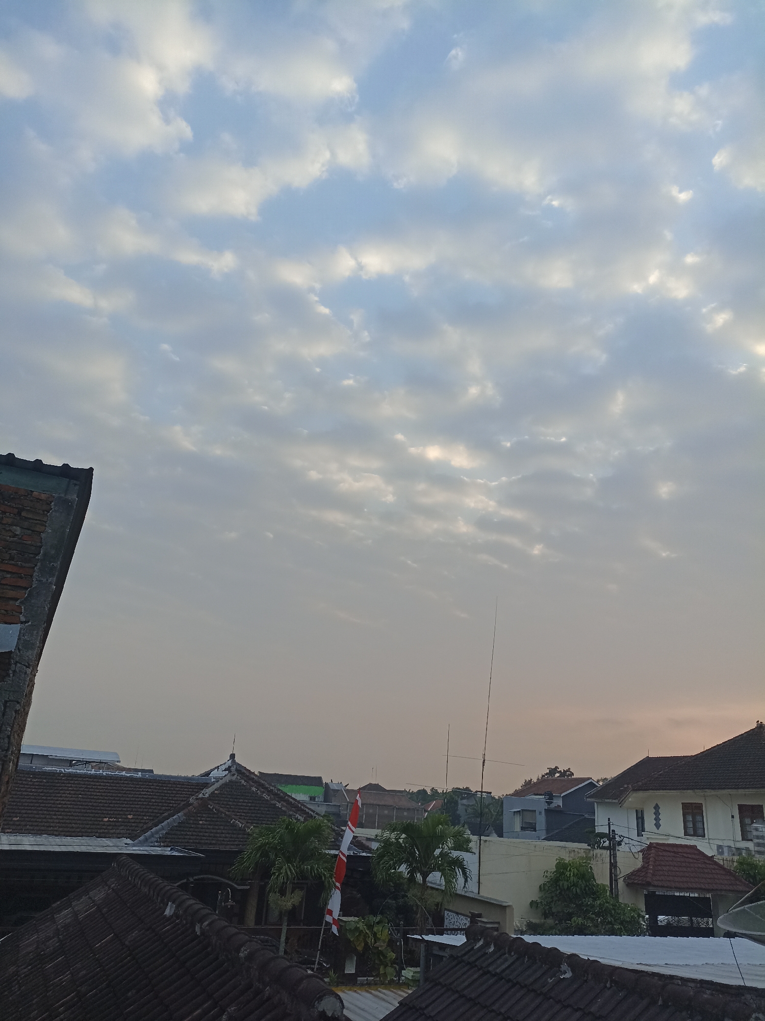 The image shows a view from a rooftop in Sumberejo, Indonesia, looking towards the sky and other rooftops. The sky is filled with scattered clouds, with patches of lighter and darker gray, suggesting it might be dawn or dusk. The atmosphere appears hazy.
In the foreground, the dark, textured tiles of several rooftops are visible. On the left side, the corner of a brick building with a rough concrete edge is present. In the mid-ground, more rooftops with dark tiles are seen, along with some palm trees. A flagpole with a red and white Indonesian flag (Sang Saka Merah Putih) is positioned between the palm trees.
Further back, various buildings with different roof styles and colors are visible, including some white houses with red roofs. Several thin, vertical antennas or poles rise from some of the buildings. The overall impression is a residential area in a town or city. No people are visible in the image.