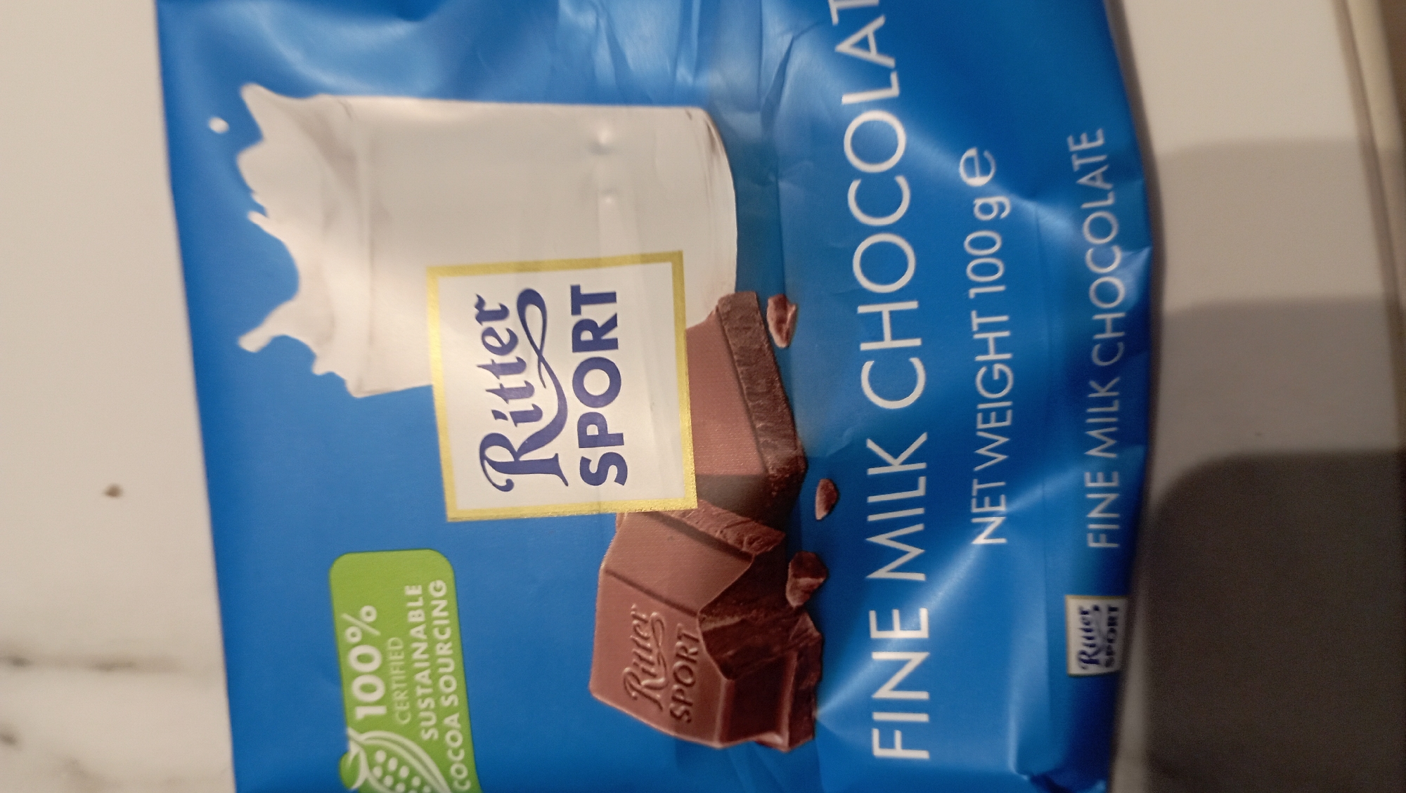 The image shows a blue Ritter Sport fine milk chocolate bar wrapper. The wrapper features the Ritter Sport logo with a splash of milk behind it, and a picture of chocolate squares. The text on the wrapper reads "FINE MILK CHOCOLAT," "NET WEIGHT 100 g e," "FINE MILK CHOCOLATE," and "100% CERTIFIED SUSTAINABLE COCOA SOURCING."