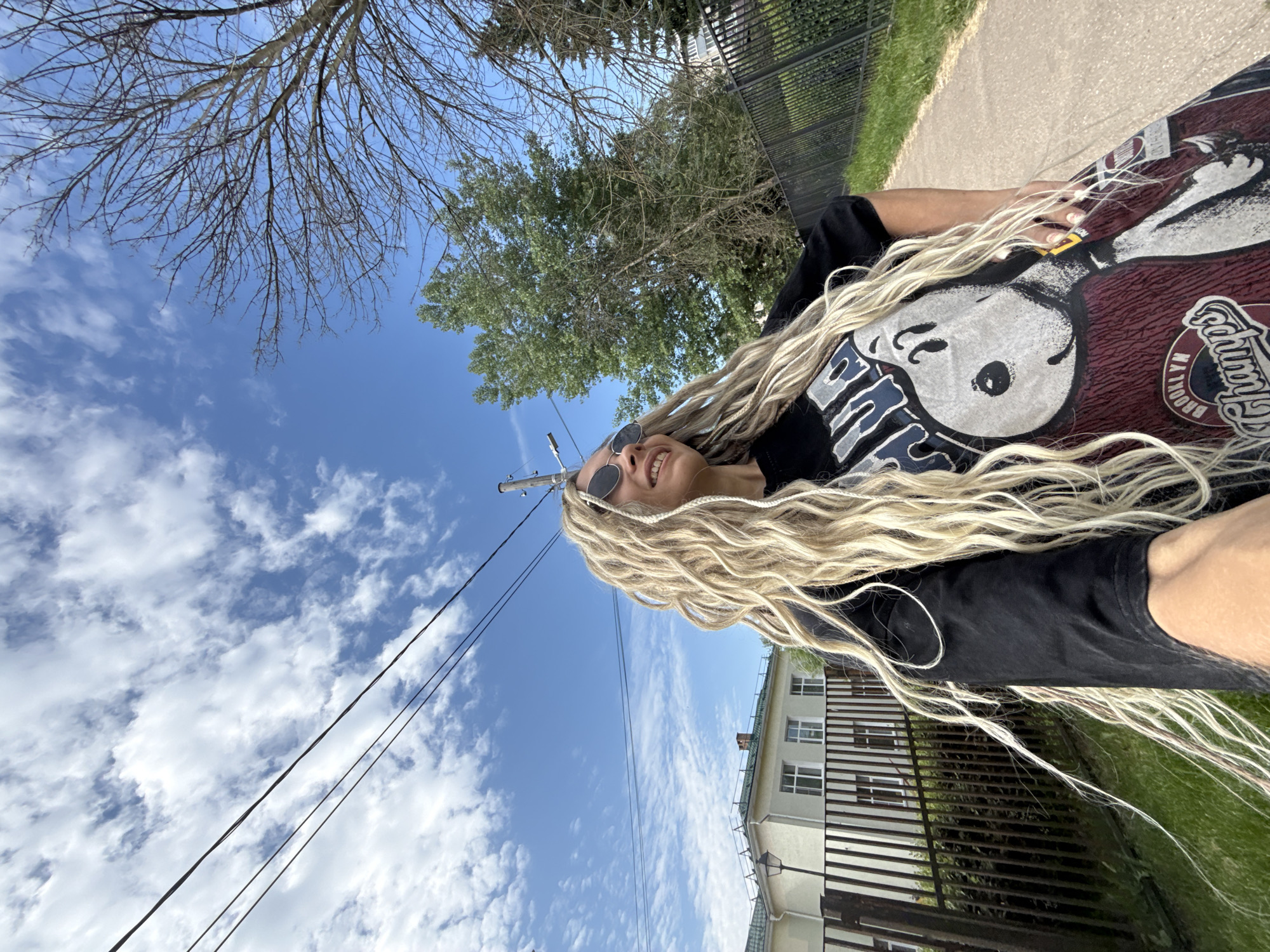 A woman with blonde dreadlocks and sunglasses is captured in a low-angle, outdoor, selfie-style shot. She is wearing a black t-shirt featuring a graphic of Snoopy and "Brooklyn Champs." The background includes a sidewalk, a dark metal fence, a light-colored building with a green roof and multiple windows, green grass, and trees. Overhead, visible power lines stretch across a bright blue sky dotted with white clouds. The location is identified as Rusanovo, Russia. The woman is smiling and looking upwards and to her right, with her right hand on her hip.