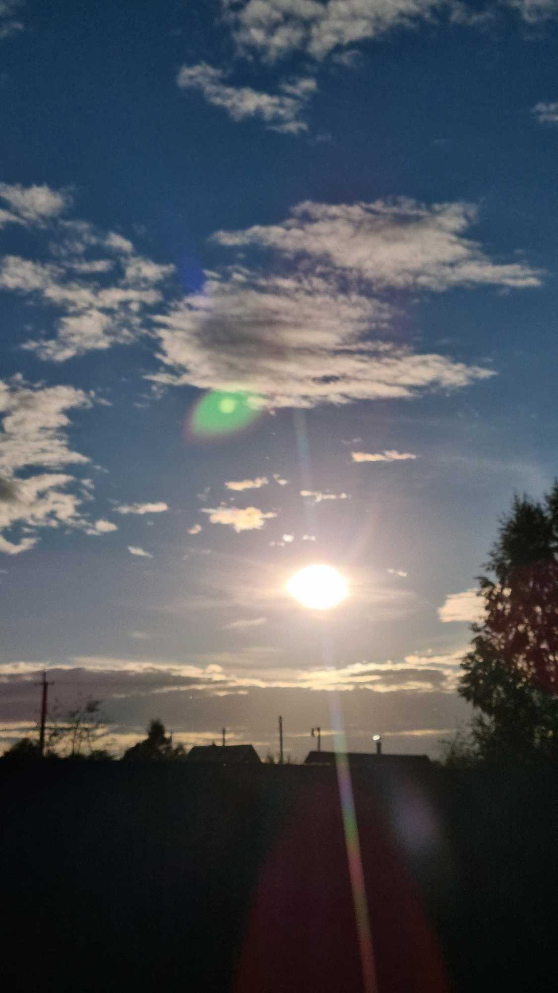 This is a vertical, medium-shot photograph of a cloudy sky at sunset, with a silhouette of houses and trees in the foreground. The sun is visible as a bright, round orb in the center of the frame, surrounded by lens flare effects that create streaks of light. Wispy white clouds are scattered across a deep blue sky. In the lower portion of the image, a dark, silhouetted landscape features the rooftops of several houses, utility poles, and the upper branches of a tree on the right. The overall scene suggests a rural or suburban setting in Vysokoe, Russia, during the evening.