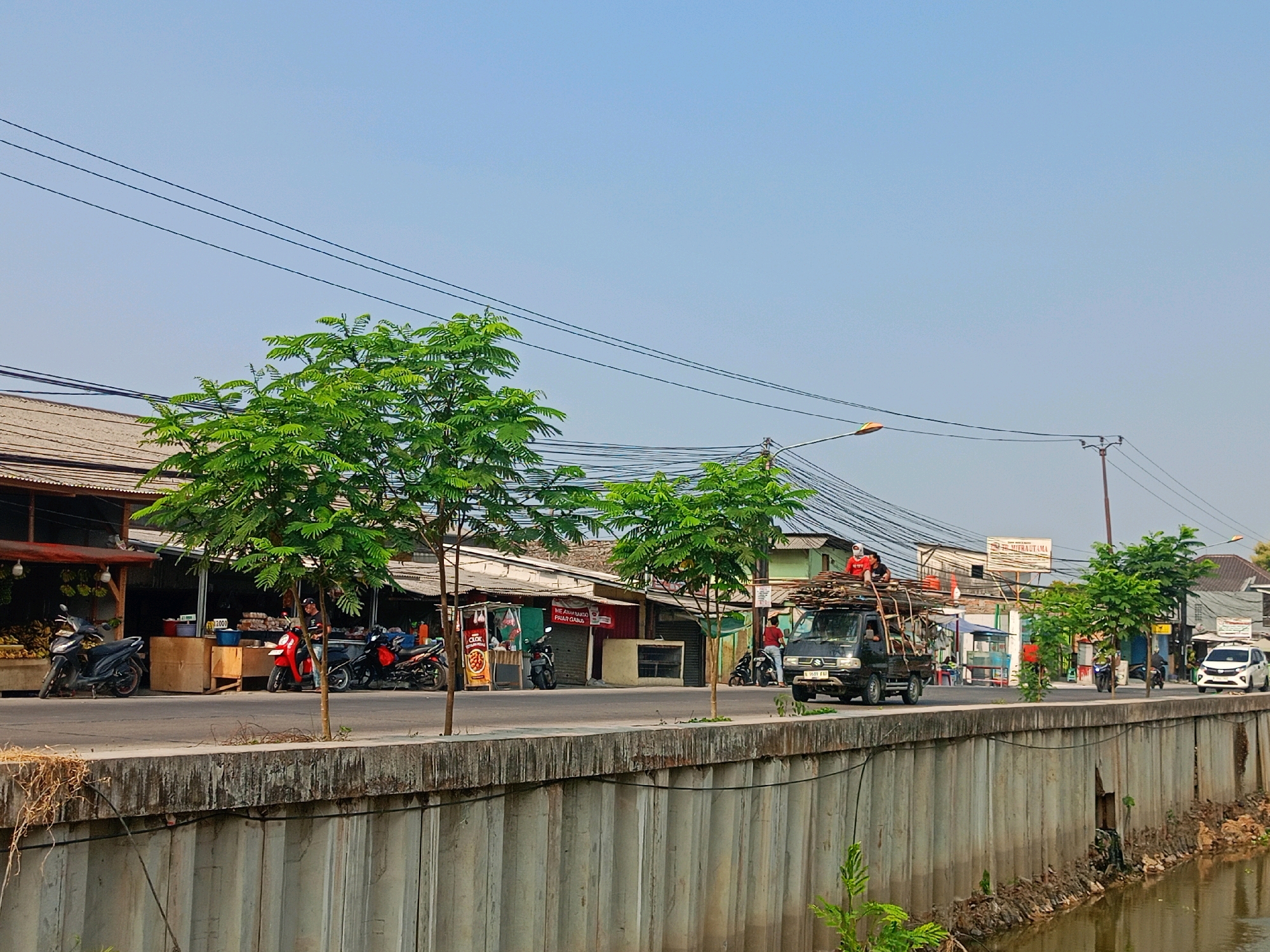 The image depicts a street scene in Sriamur, Indonesia. A paved road runs horizontally across the frame, bordered by a concrete wall on the right which overlooks a body of water. On the left side of the road, a row of small shops with corrugated roofs are visible. Several motorcycles are parked in front of these shops, and one red motorcycle has a person standing next to it. Two trees with green foliage are planted along the roadside.
A black pickup truck loaded with construction materials, such as rebar and wood, is traveling on the road from left to right. Two individuals are visible on top of the load of materials in the truck. Further down the road, a white SUV and a motorcycle are also in motion. The sky is clear and blue, indicating a sunny day. Electrical wires crisscross above the street and shops. The overall impression is of a busy, everyday scene in a populated area.