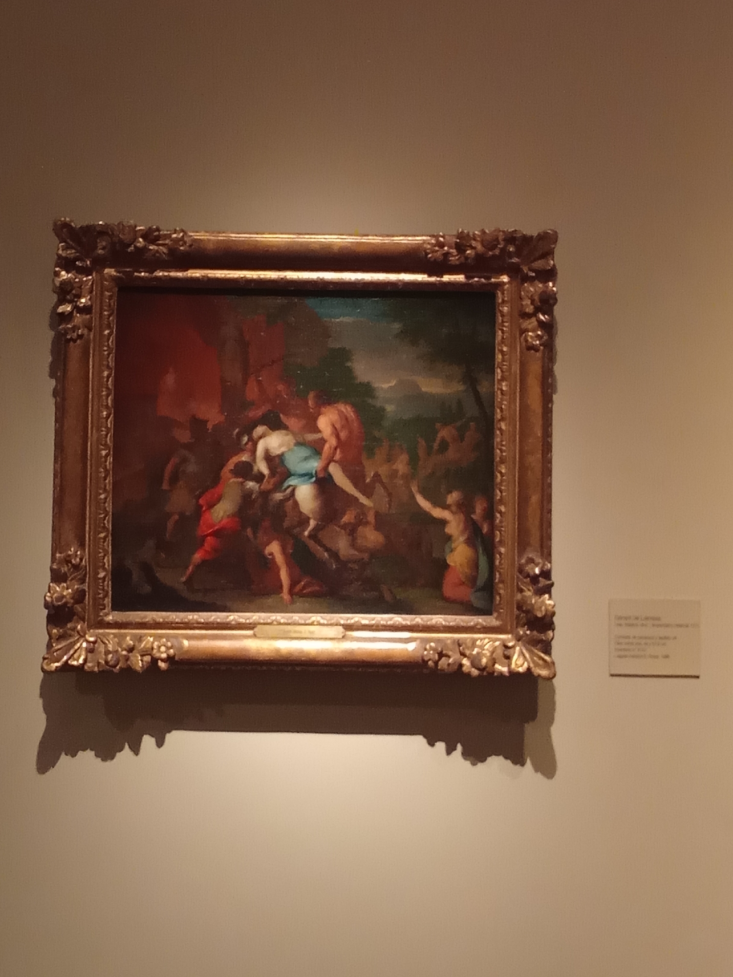 The image shows a framed oil painting displayed on a beige wall. The painting depicts a scene with several nude or partially clothed figures in a dynamic, possibly chaotic, composition. The figures appear to be engaged in some form of struggle or movement. The background of the painting includes elements of landscape, with greenery on the right and a red, possibly fiery, element on the left. The painting is enclosed in an ornate, gold-colored frame with decorative carvings.
To the right of the painting, a small, rectangular label with text is affixed to the wall. The text on the label is mostly illegible due to the image quality and distance, but it appears to be an exhibit or artwork description.
The overall setting suggests an indoor exhibition space, likely a museum or gallery. Based on the presence of a label with Spanish text and the style of art commonly exhibited in prominent Buenos Aires institutions, this artwork is likely located at the Museo Nacional de Bellas Artes (MNBA) in Buenos Aires, Argentina. The museum is known for its extensive collection of Argentine and international art.