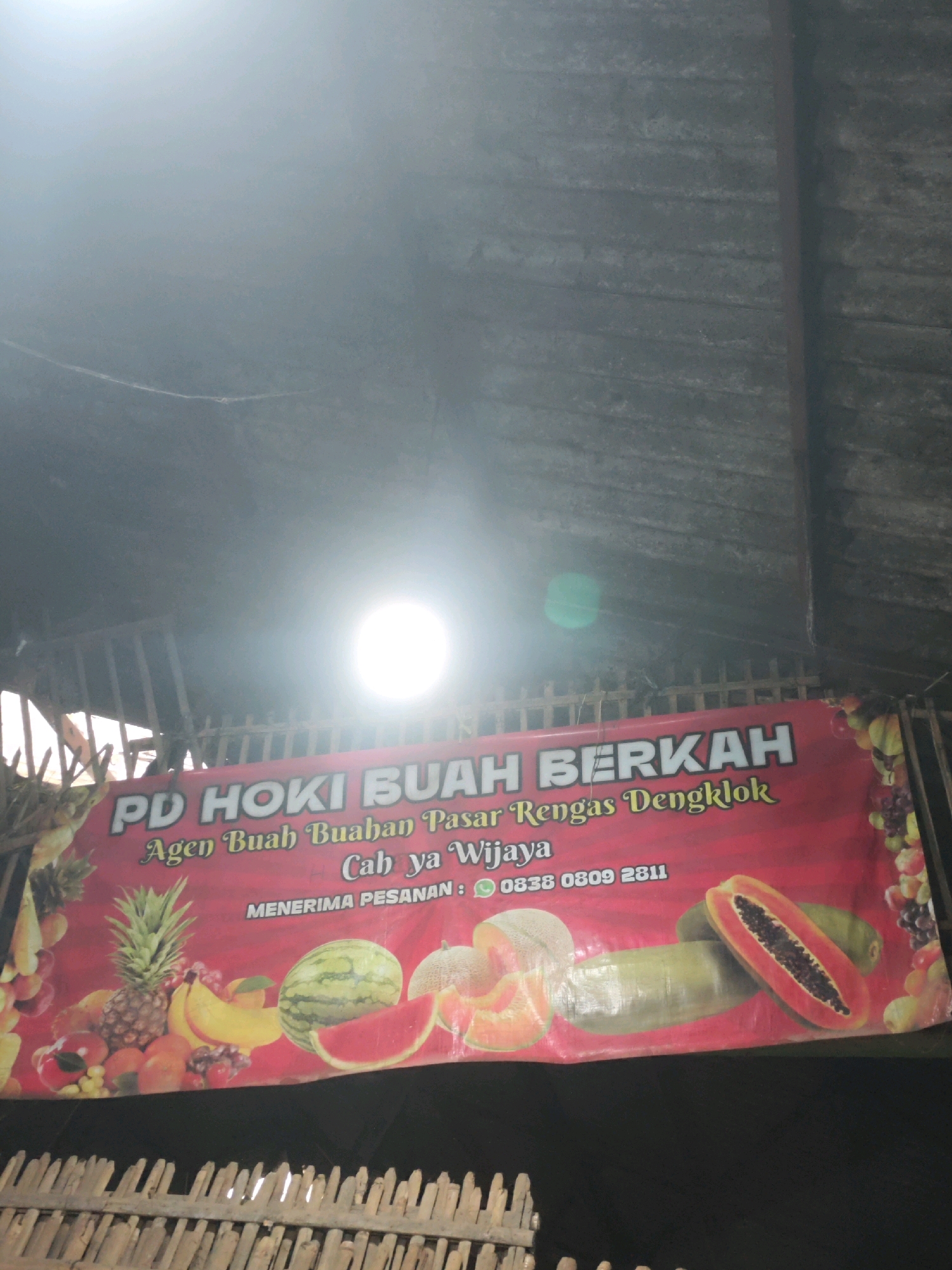 This image shows a banner for "PD HOKI BUAH BERKAH," an agent for fruits in Rengas Dengklok Market, with the branch located in Wijaya. The banner displays various fruits like pineapple, bananas, watermelon, papaya, and melon, along with text indicating it accepts orders. Below the banner, a wooden structure is visible. The banner is hung outdoors, likely in an area with a covered roof, suggesting it is a stall or shop. The lighting and background suggest it is daytime, with the sun creating a bright glare. Rengas Dengklok is a subdistrict within the Karawang Regency in West Java, Indonesia.