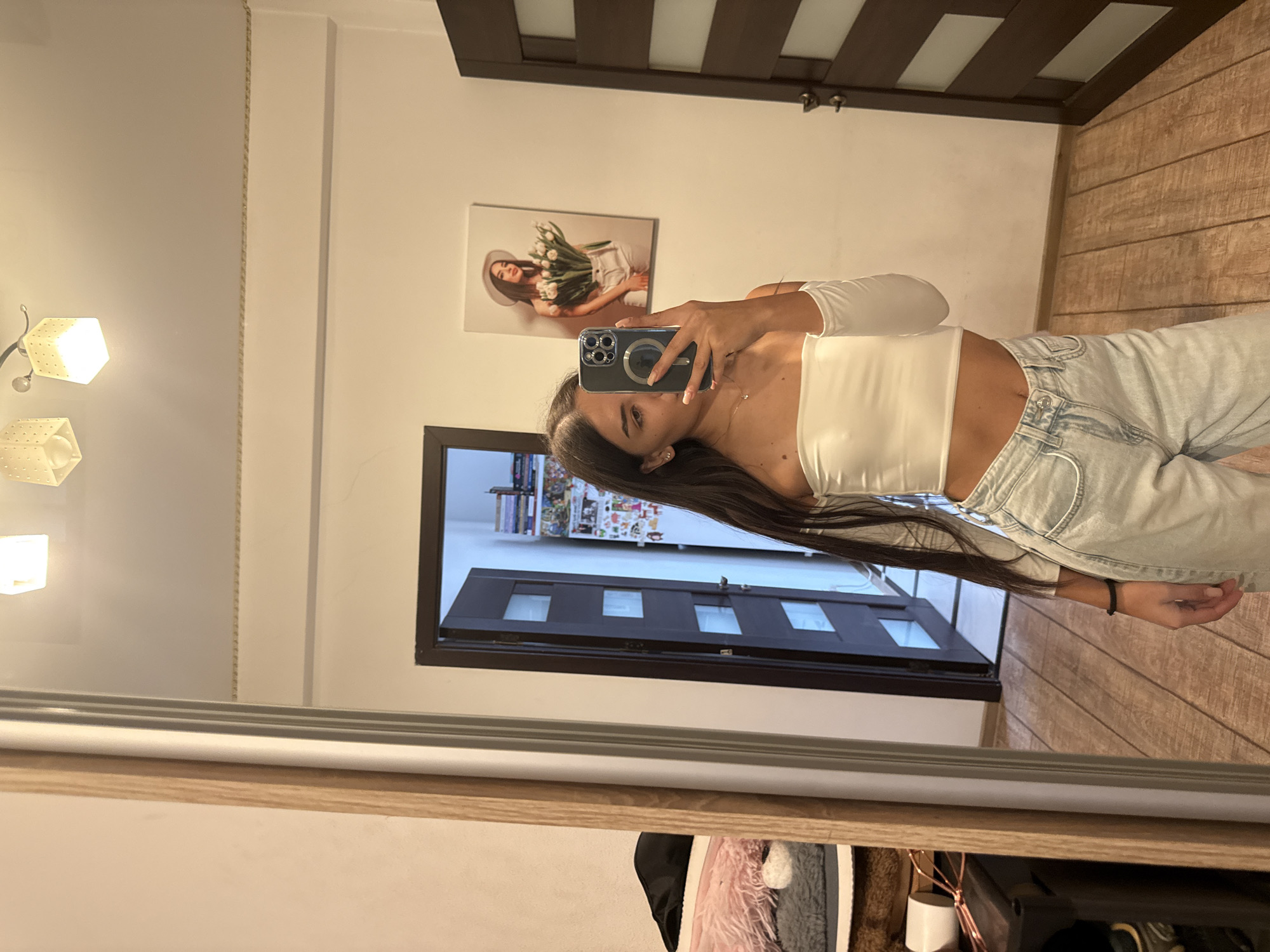A young woman is taking a mirror selfie in what appears to be a bedroom. She is wearing a white off-the-shoulder top and light-wash jeans. The room has white walls, a wooden floor, and a dark wooden door. On the wall, there is a framed picture of a woman holding tulips and wearing a hat. To the left of the frame, a large mirror reflects the scene. Behind the mirror, some furniture and items are visible, including a stack of books and a fluffy pink object. The overall impression is of a casual, personal space. There is no specific information to indicate this location is in Chișinău, Moldova.