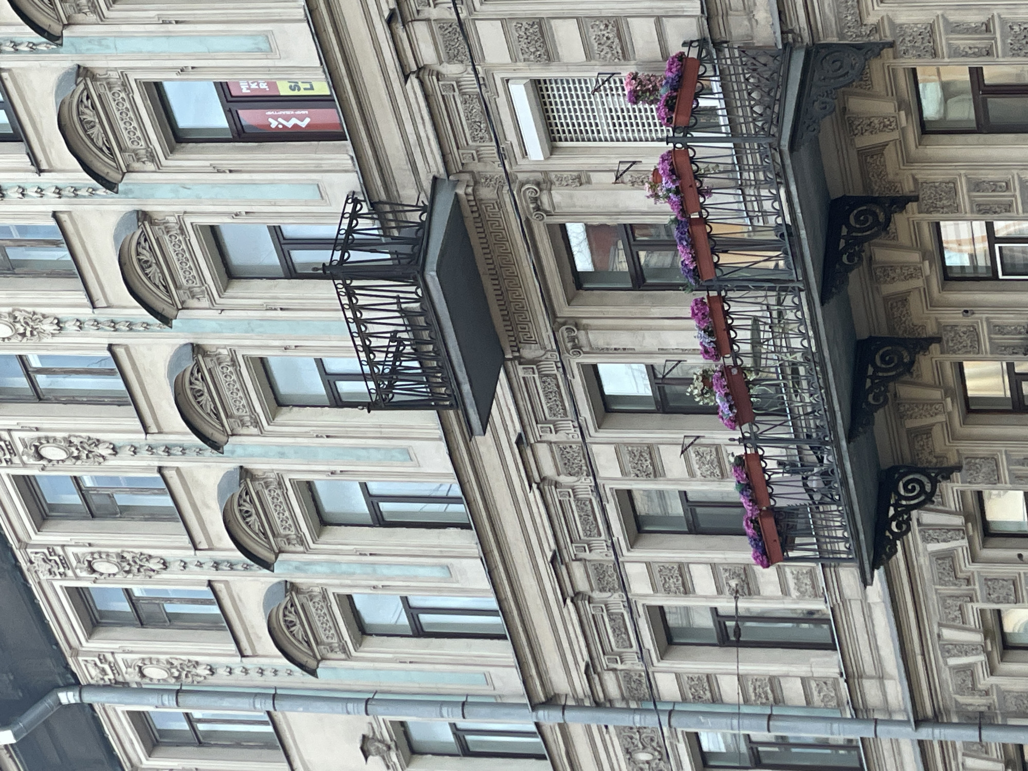 The image shows a section of a building's facade, featuring multiple windows and ornate balconies. The architecture is consistent with that found in Saint Petersburg, Russia, characterized by decorative embellishments and historical style. The balconies are adorned with flower boxes containing purple flowers. The facade exhibits a gray-beige color palette. A pipe runs vertically along the side of the building. The architectural details suggest a historic structure.