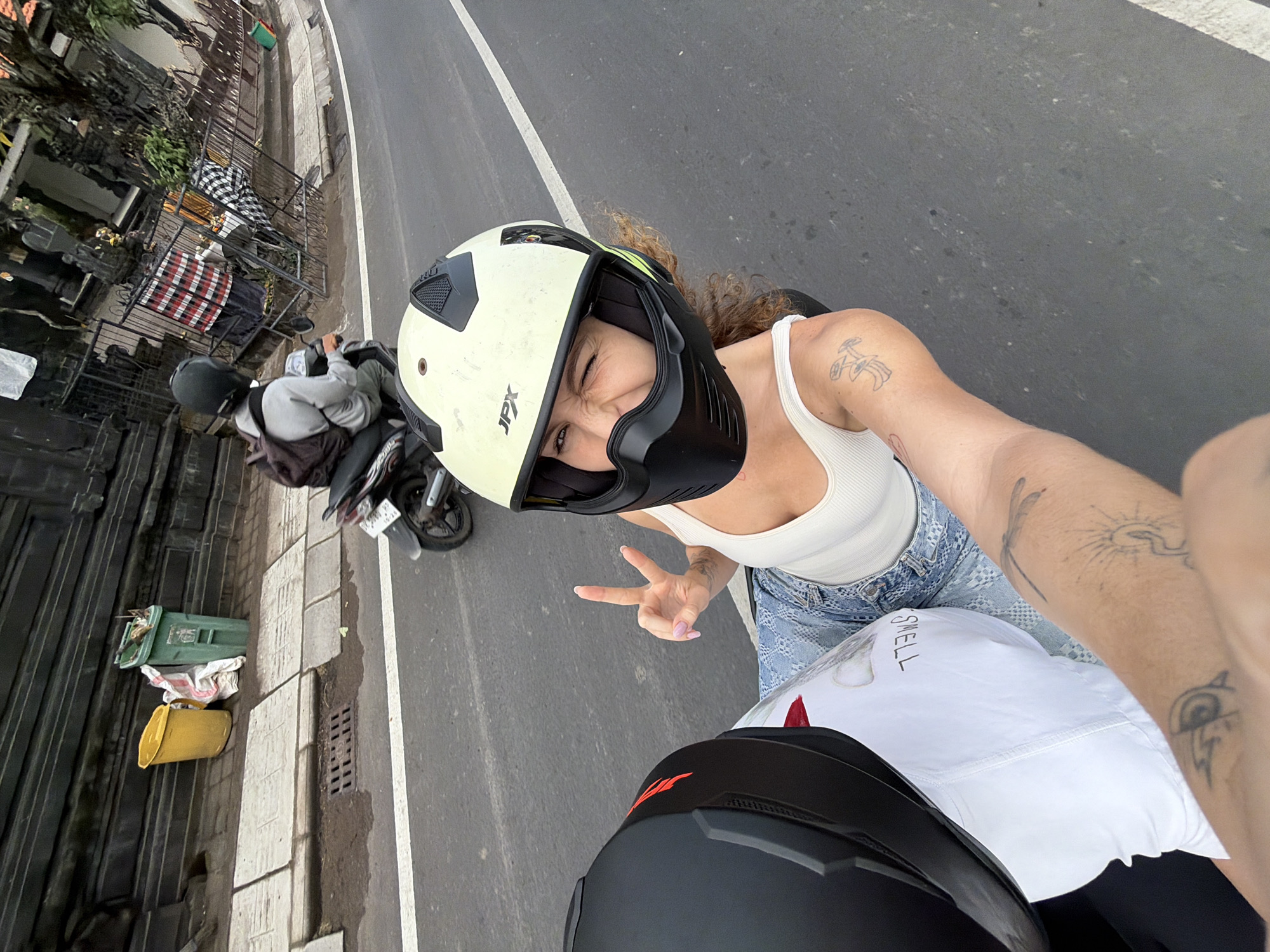 In the city of Mengwi, Indonesia, a person with curly hair, wearing a white tank top and blue denim jeans, is riding on the back of a motorcycle, capturing a selfie. They are wearing a light-colored helmet with a black face shield and making a peace sign with their right hand. The person in front, wearing a black helmet, is mostly obscured by the camera angle. The motorcycle is traveling on a paved road, indicated by the white line marking the lane. In the background, a person on another motorcycle is visible, along with a building with intricate dark stonework, a black metal fence, and traditional Balinese checkered fabrics. Yellow and green trash bins are also present on the roadside. The overall scene depicts a moment of travel and exploration in an urban Indonesian setting.