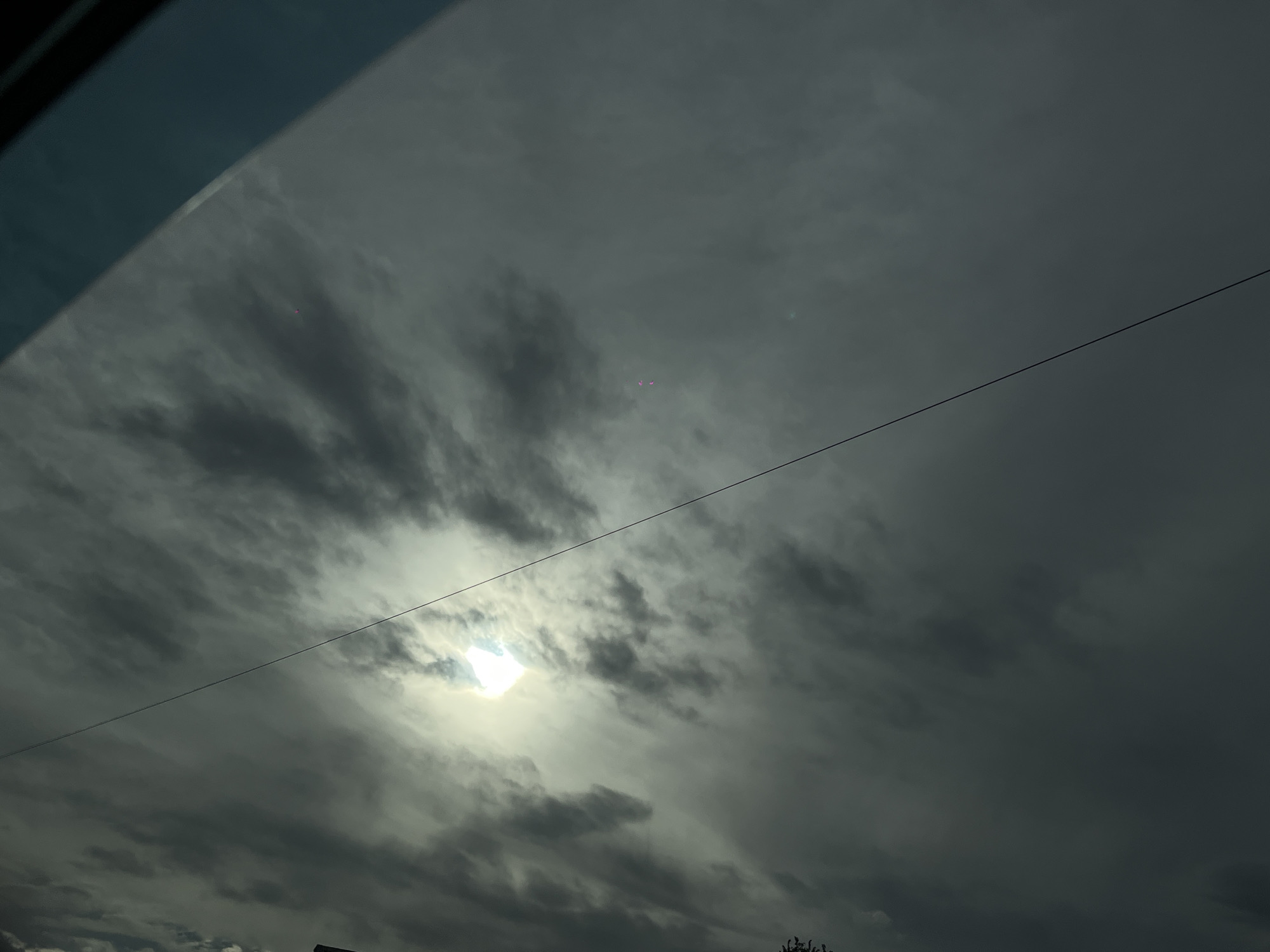 The image shows a sky view, presumably from Novopokrovskaya, Russia, characterized by heavy cloud cover. The sun is visible through a thin layer of clouds, creating a bright spot. A power line runs diagonally across the frame. The bottom of the image shows the tops of trees or shrubbery. In the upper left corner, a portion of the interior of a vehicle (possibly a car roof) is visible. Two red spots are barely visible in the center of the image.