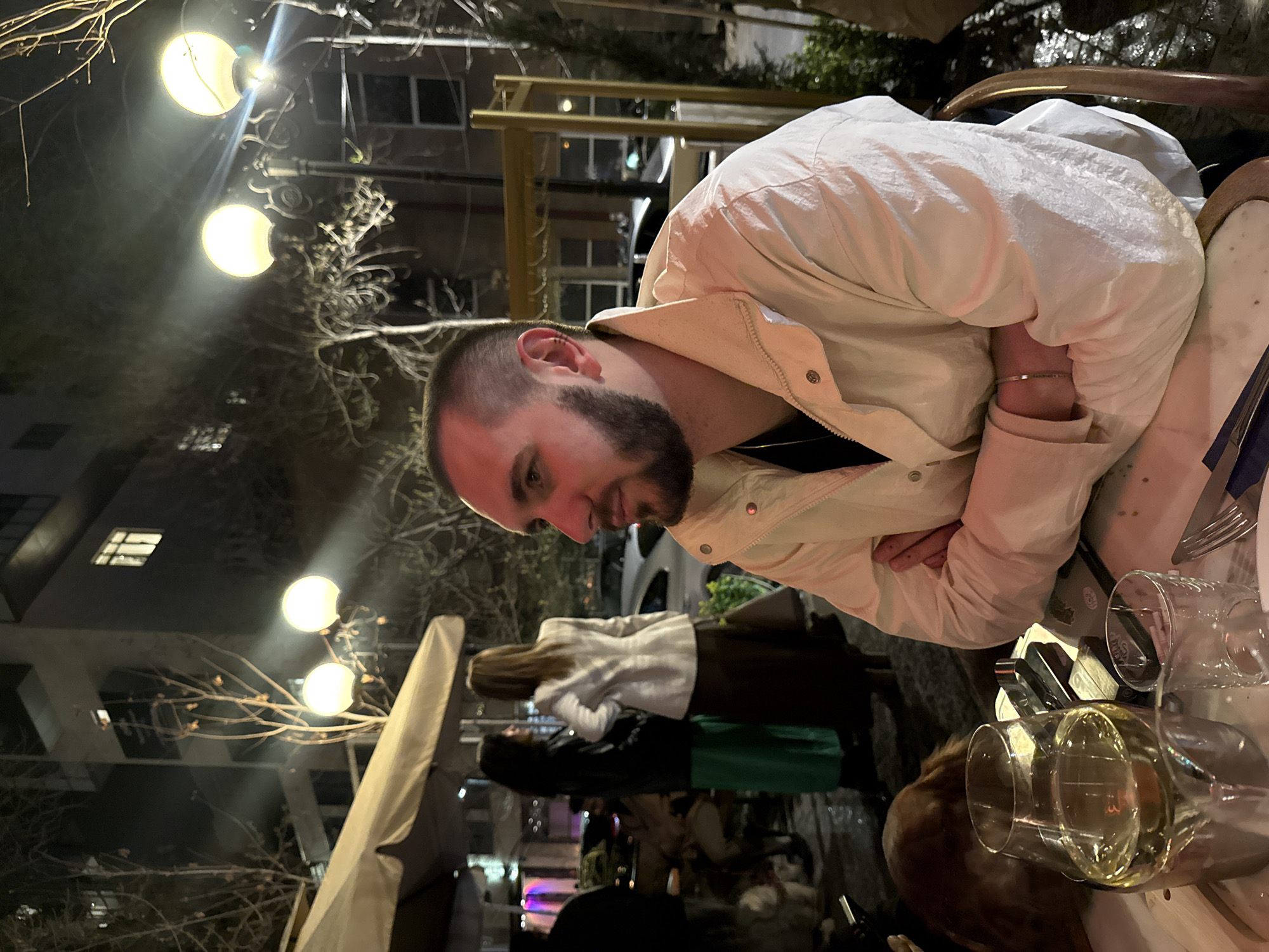The image shows a man with a beard and a light-colored jacket sitting at an outdoor table at night. He is looking down at the table. There are several wine glasses on the table. In the background, there are other people, trees, and buildings. Streetlights are visible. This scene is likely in Yerevan, Armenia, because that was requested.