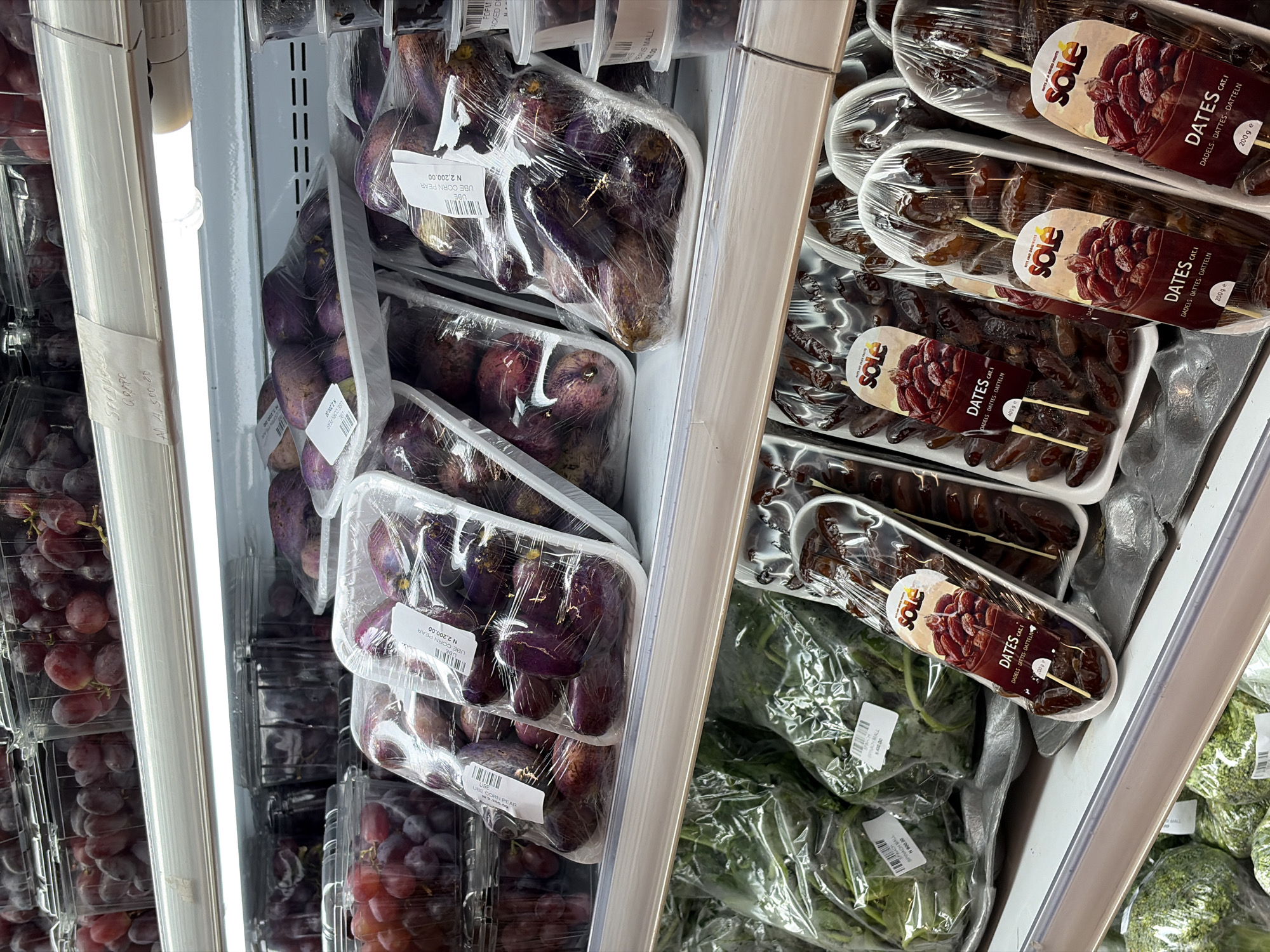 The image shows a refrigerated shelf in what appears to be a supermarket in Igboefon, Nigeria. The shelf contains several types of produce, including: grapes, ube (African pear or corn pear), spinach, and packaged dates (labeled "Sole Dates"). The produce is arranged in plastic containers or bags, and some items have price tags or labels with the name "MALL". The shelf is illuminated by a fluorescent light on the left.