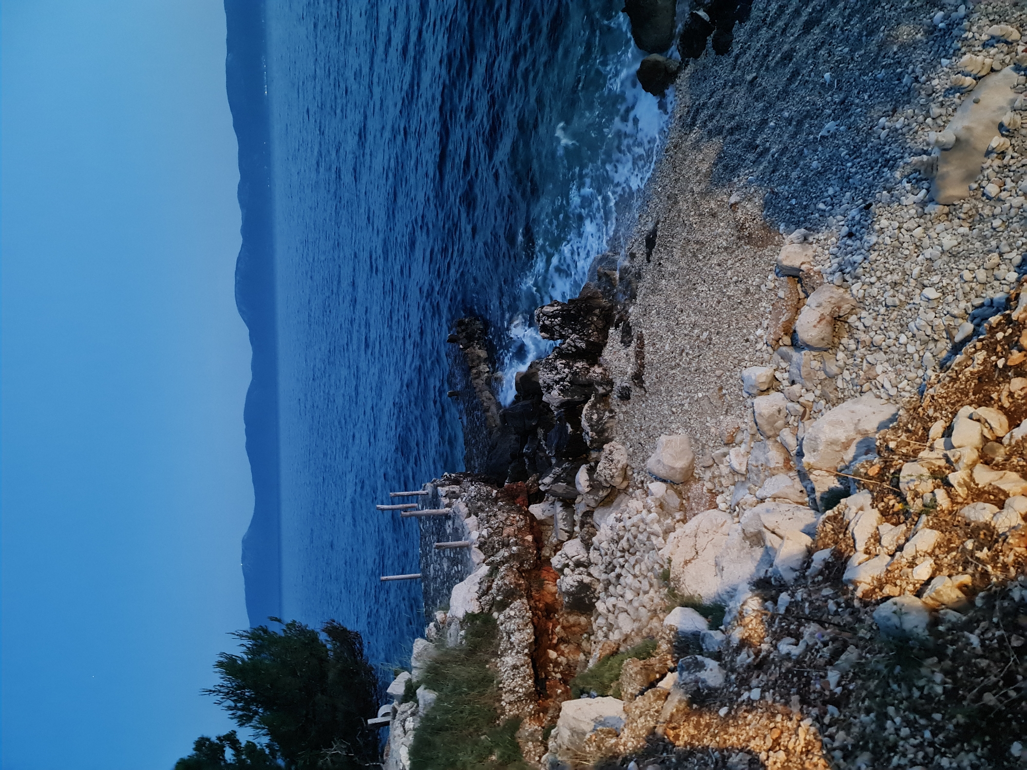 The image depicts a rocky coastline at dusk or dawn, likely in Brist, Croatia. The scene is dominated by a vast expanse of deep blue sea stretching to the horizon, where distant, hazy mountains are visible. The shoreline is composed of numerous small, smooth pebbles and larger, rougher rocks.
In the foreground, a section of the rocky shore is illuminated by warm, artificial light, creating a stark contrast with the cooler tones of the sea and sky. A small, weathered wooden structure, possibly the remains of a pier or jetty, juts out into the water from the rocky embankment on the left side of the image. This structure consists of a flat, irregular stone platform with several vertical wooden posts rising from it.
Vegetation, including a dark green pine tree, grows on the elevated rocky area to the left. The water shows gentle ripples and a small amount of white foam along the shore, indicating the presence of waves. The sky is a clear, deep blue, with a faint hint of lighter blue near the horizon. No people are visible in the image. The overall impression is one of a quiet, natural coastal setting.