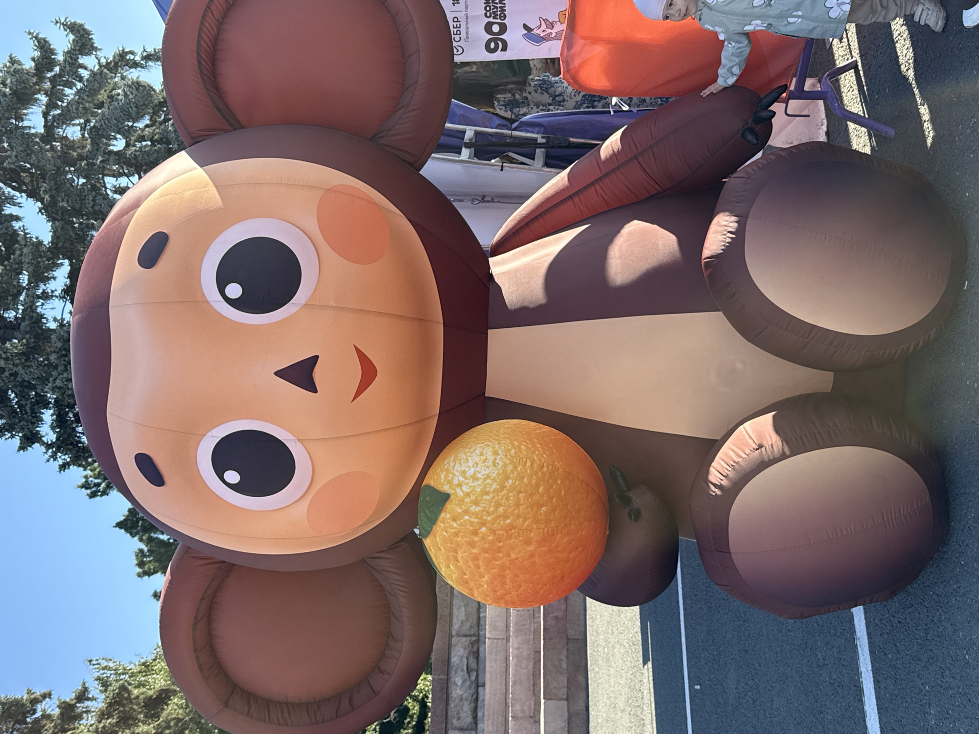 A large, inflatable character resembling Cheburashka, a beloved Soviet-era cartoon character, stands outdoors, holding a bright orange, inflated sphere that resembles a tangerine. The character is brown and tan, with large, round ears and big, black eyes. A young child, wearing a light-colored jacket with a floral pattern and a white hat, stands to the right of the inflatable, reaching out towards it. Behind the inflatable, a banner with Russian text and logos, including one for Sberbank, is visible. The scene appears to be set on an asphalt surface, possibly a city square or park, with trees and a blue sky in the background. The presence of the Sberbank banner and the likely context of an event suggest this image was taken in Chelyabinsk, Russia, possibly during a public celebration or festival.