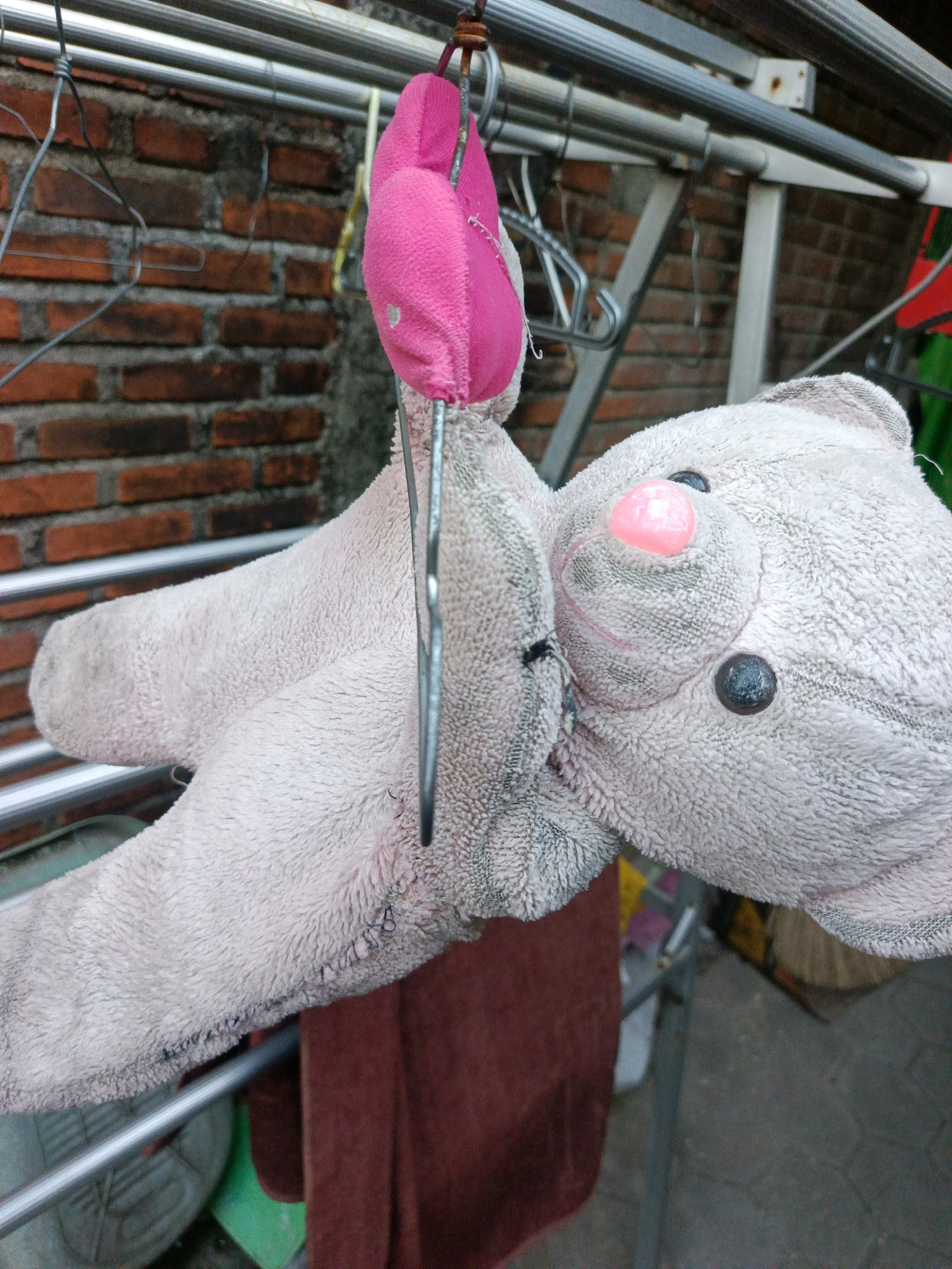 A close-up, eye-level outdoor shot shows a light pink stuffed bear hanging from a metal clothesline, its body draped over a brown towel. The bear is positioned so its face, featuring a pink nose and dark eyes, is visible. The background consists of a red brick wall and a metal drying rack. The scene is likely located in Kwangsan, Indonesia.