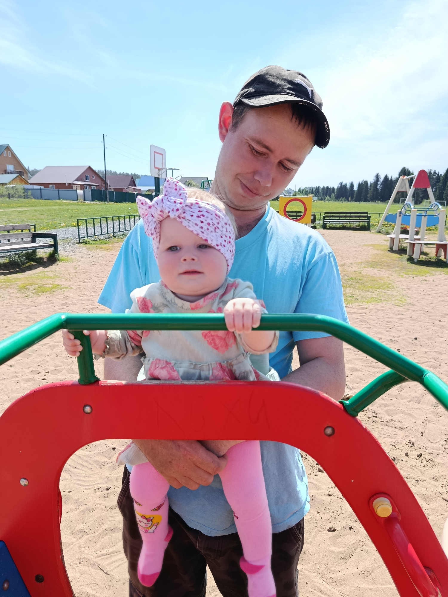 On a sunny day in Фоки, Russia, a man wearing a black baseball cap and a light blue t-shirt is holding a baby with a pink and white polka-dotted headband. The baby wears a light patterned dress and pink tights with cartoon characters. They are standing in a sandbox next to a red and green playground structure. In the background, there is a basketball hoop, houses, and a grassy area.
