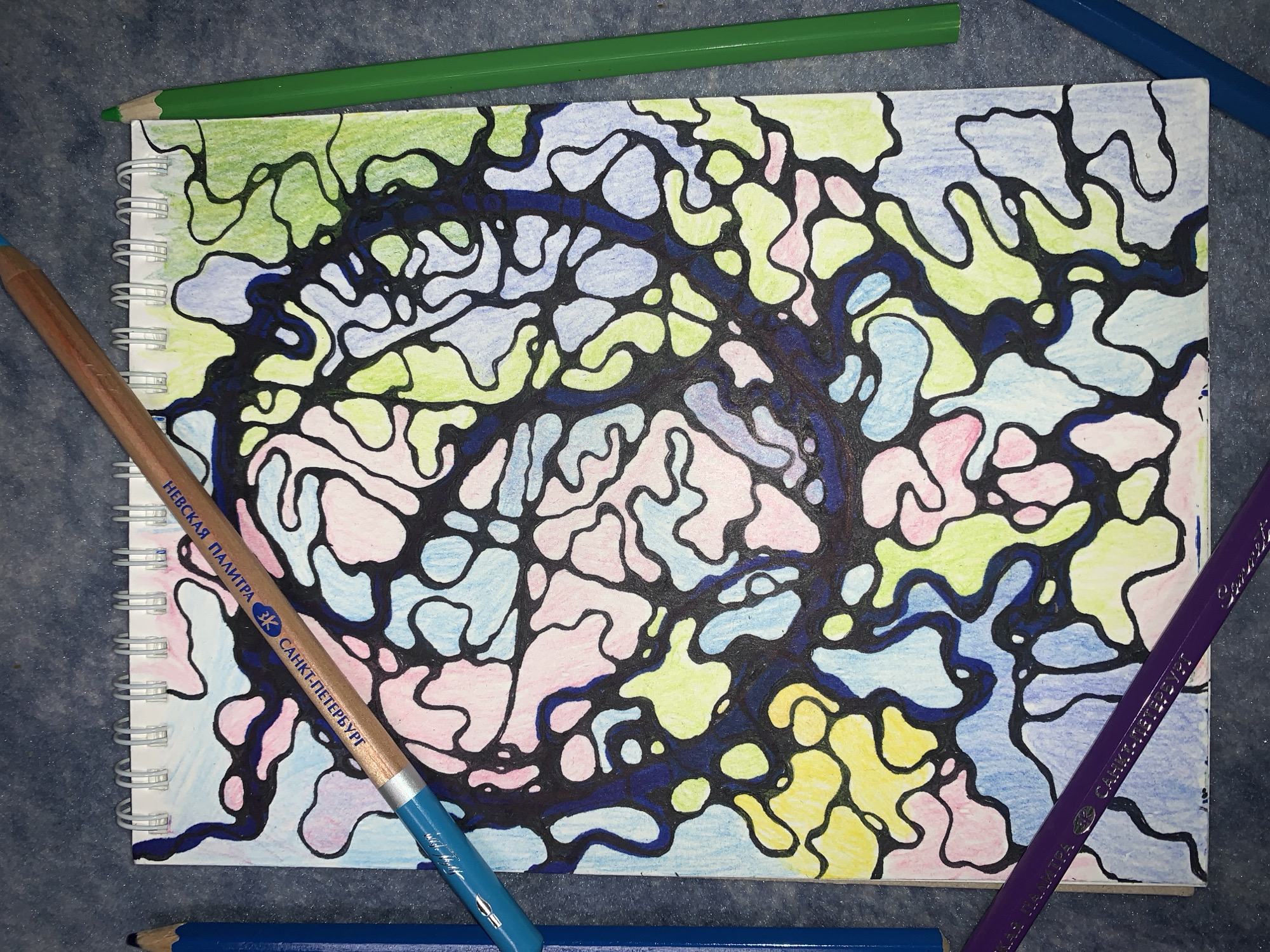 A close-up, overhead shot shows a drawing on a spiral-bound notebook. The drawing is abstract, with black, irregular shapes that create cells of color. The colors filling the cells are light green, yellow, pink, and blue. A wooden pencil with blue lead is placed diagonally across the bottom right of the notebook. It has text printed on it in Russian: "НЕВСКАЯ ПАЛИТРА" and "САНКТ-ПЕТЕРБУРГ". A green colored pencil lies across the top of the notebook, and a purple colored pencil is at the bottom right, diagonally across the drawing. A blue pencil is visible at the bottom edge of the frame. The drawing appears to be a neurographic art piece, which is a technique used for stress relief and self-exploration. The background is a dark gray, textured surface, possibly a blanket or a table. There are no people, or any indication of time of day, weather, or specific location. The overall emotion is one of calm and creativity.