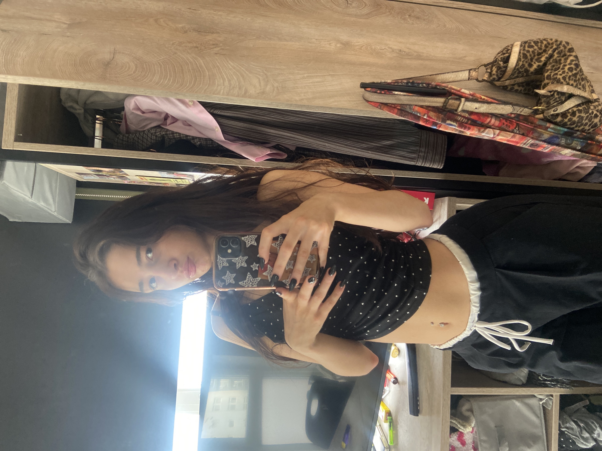 A young adult female is depicted taking a mirror selfie indoors. She possesses long, dark hair and wears a black crop top with white polka dots, revealing her midriff which features a navel piercing. She also wears black trousers with a white drawstring waistband. Her fingernails are painted black, and she holds a smartphone with a star-patterned case in both hands, positioned to capture her reflection. Her facial expression includes pursed lips.
The immediate environment visible behind her includes a light wood-grain wardrobe on the right, with a section partially open to reveal clothes on hangers, including a pink shirt and a striped shirt. A leopard-print handbag with gold accents and a patterned strap are suspended from the side of the wardrobe. To the left, a light wood-grain desk surface holds a large, black television or monitor, which reflects an exterior building with multiple windows, illuminated by a bright light source. On the desk, multiple small items are visible, including a red and white cigarette pack, lighters, and pens. Below the desk, storage cubbies contain various soft materials.
The scene provides no explicit visual markers to identify a specific location within London, United Kingdom.